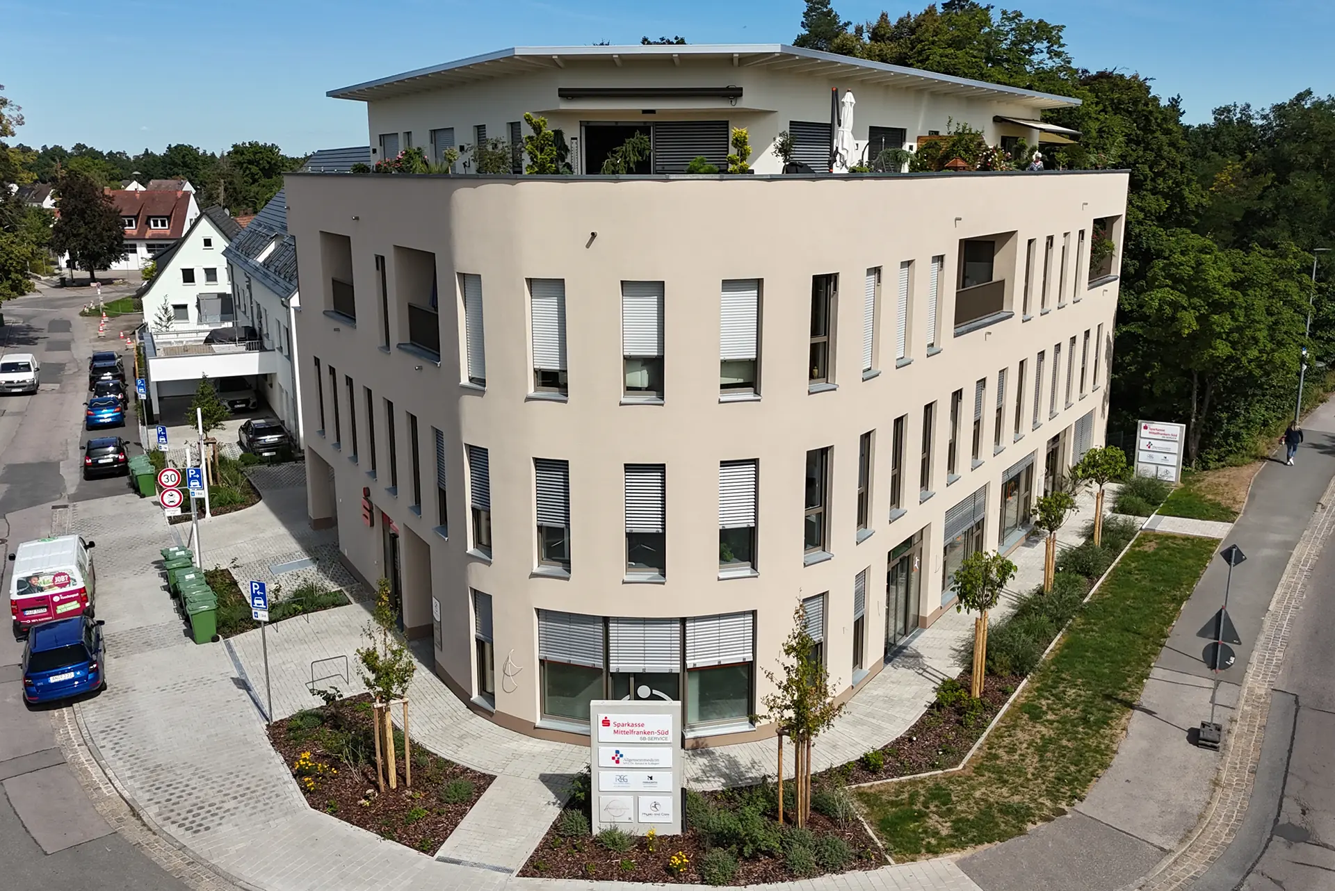 Modernes, dreigeschossiges Bürogebäude mit hellbeiger Fassade, großen Fenstern und begrüntem Dach. Umgeben von einer gepflasterten Fläche, Bäumen und Parkplätzen.