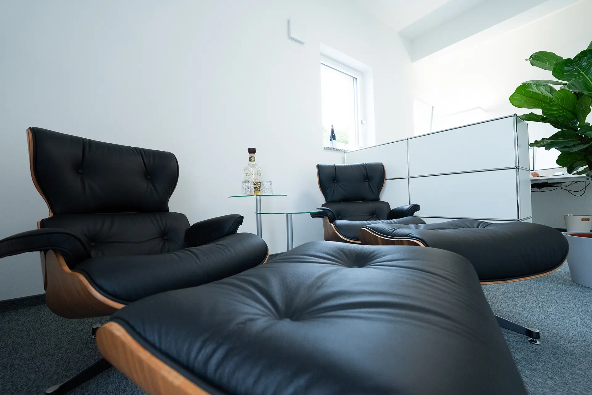 Zwei schwarze Leder-Sessel mit Holzrahmen stehen in einem Raum. Ein kleiner Glastisch befindet sich zwischen den Sesseln. Im Hintergrund ist eine Pflanze und ein Schreibtisch sichtbar.
