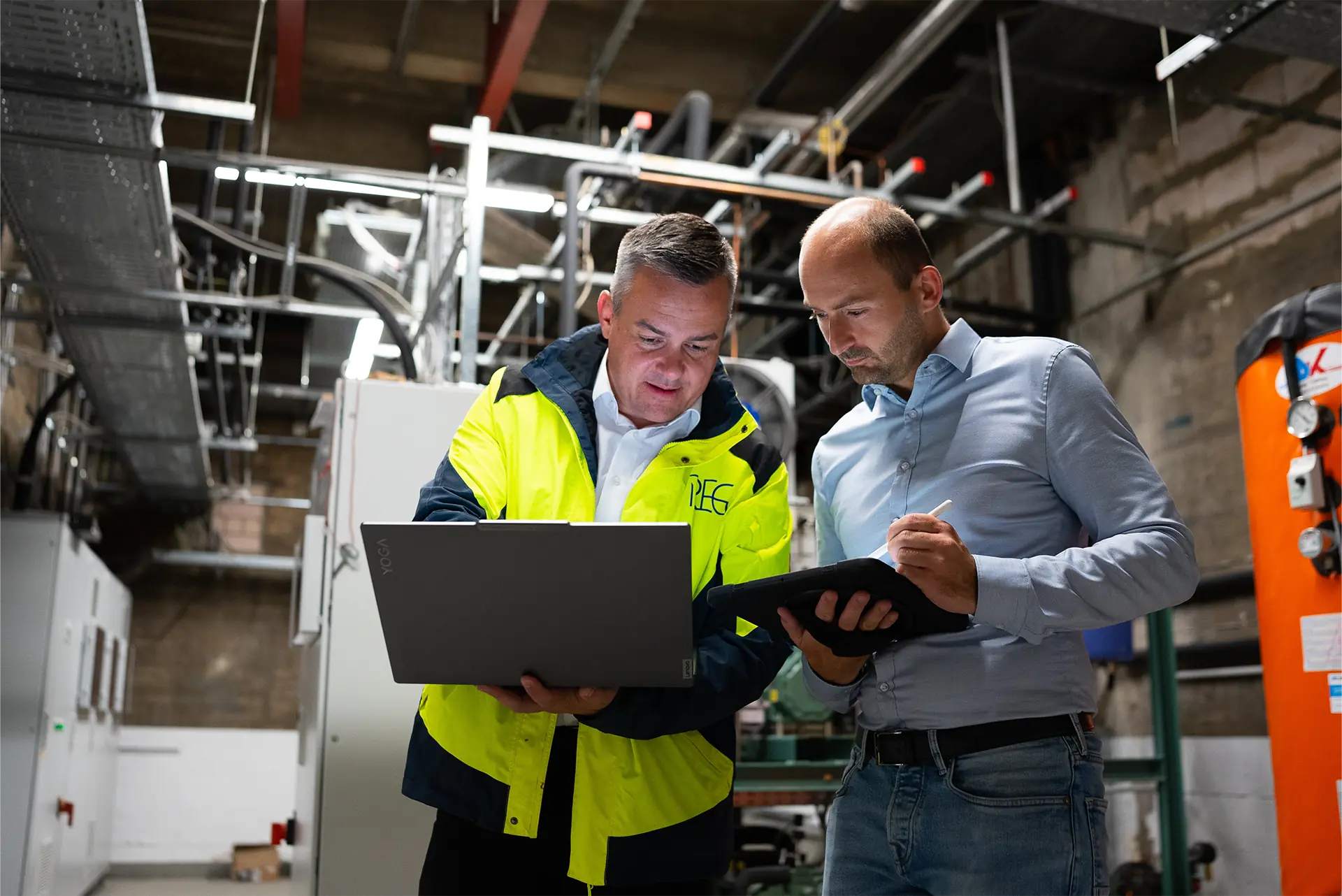 Zwei Männer in Arbeitskleidung und Hemd stehen in einer Industrieanlage, einer hält Laptop, der andere ein Tablet.