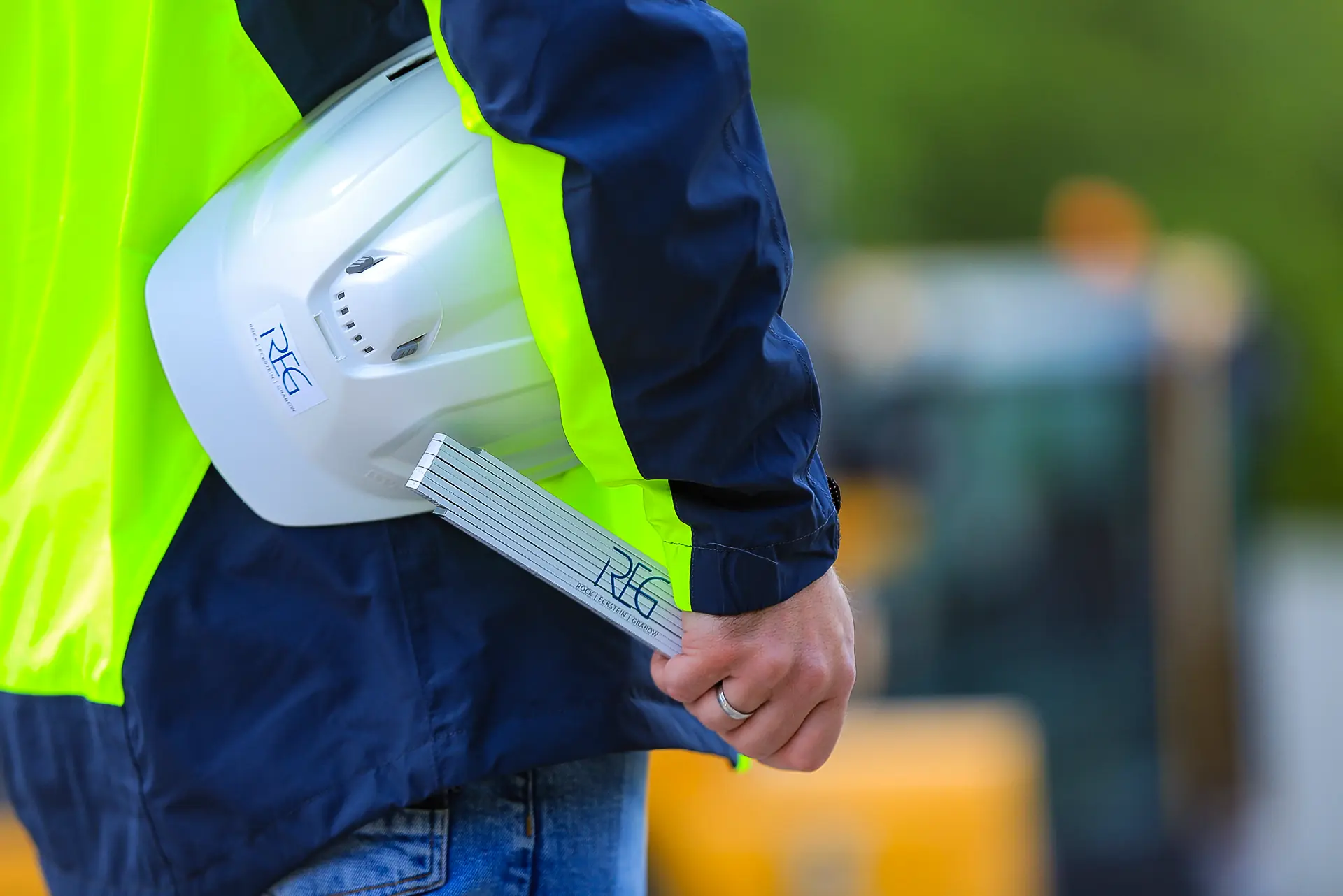 Eine Person trägt eine gelbe Warnjacke und hält einen weißen Helm unter dem rechten Arm geklemmt. In der rechten Hand hält sie einen Meterstab. Im Hintergrund sind unscharfe Baustellenfahrzeuge zu sehen.