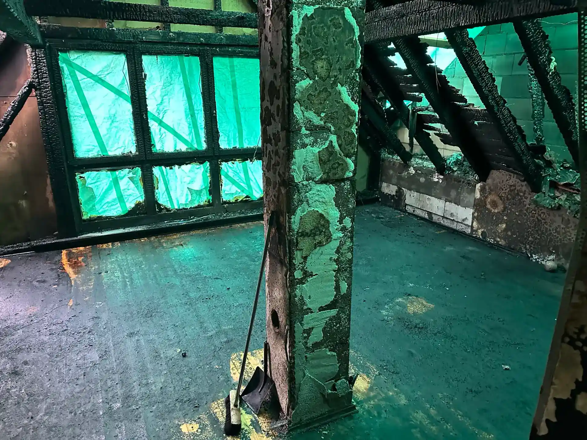 Zerstörter Dachboden mit verkohlten Holzbalken, Boden mit Brandrückständen bedeckt. Eine grüne Abdeckung ist durch die zerstörten Fenster und das teilweise offene Dach zu sehen.