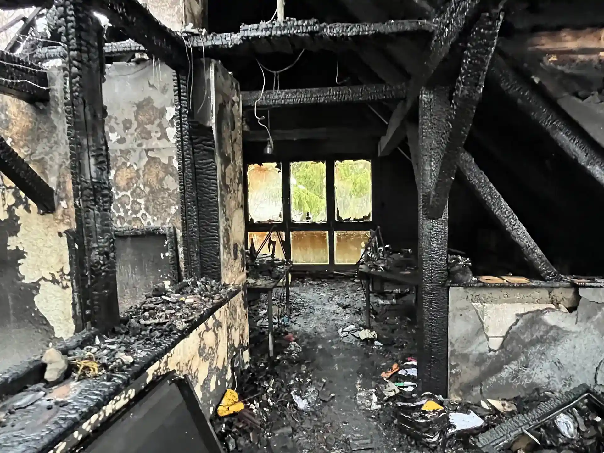 Zerstörter Dachboden nach Brandschaden. Die Holzbalken sind stark verkohlt, das Dach ist teilweise eingestürzt und die Fenster zerborsten. Auf dem Boden liegen Schutt und Asche.