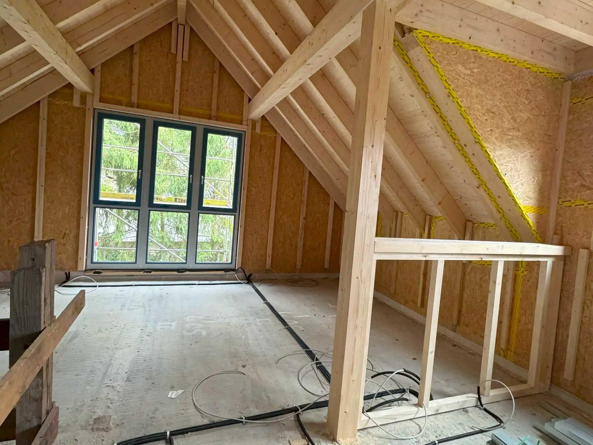 Innenraum eines Dachgeschosses mit sichtbaren Holzbalken und OSB-Platten, Kabel liegen auf dem Boden verteilt.