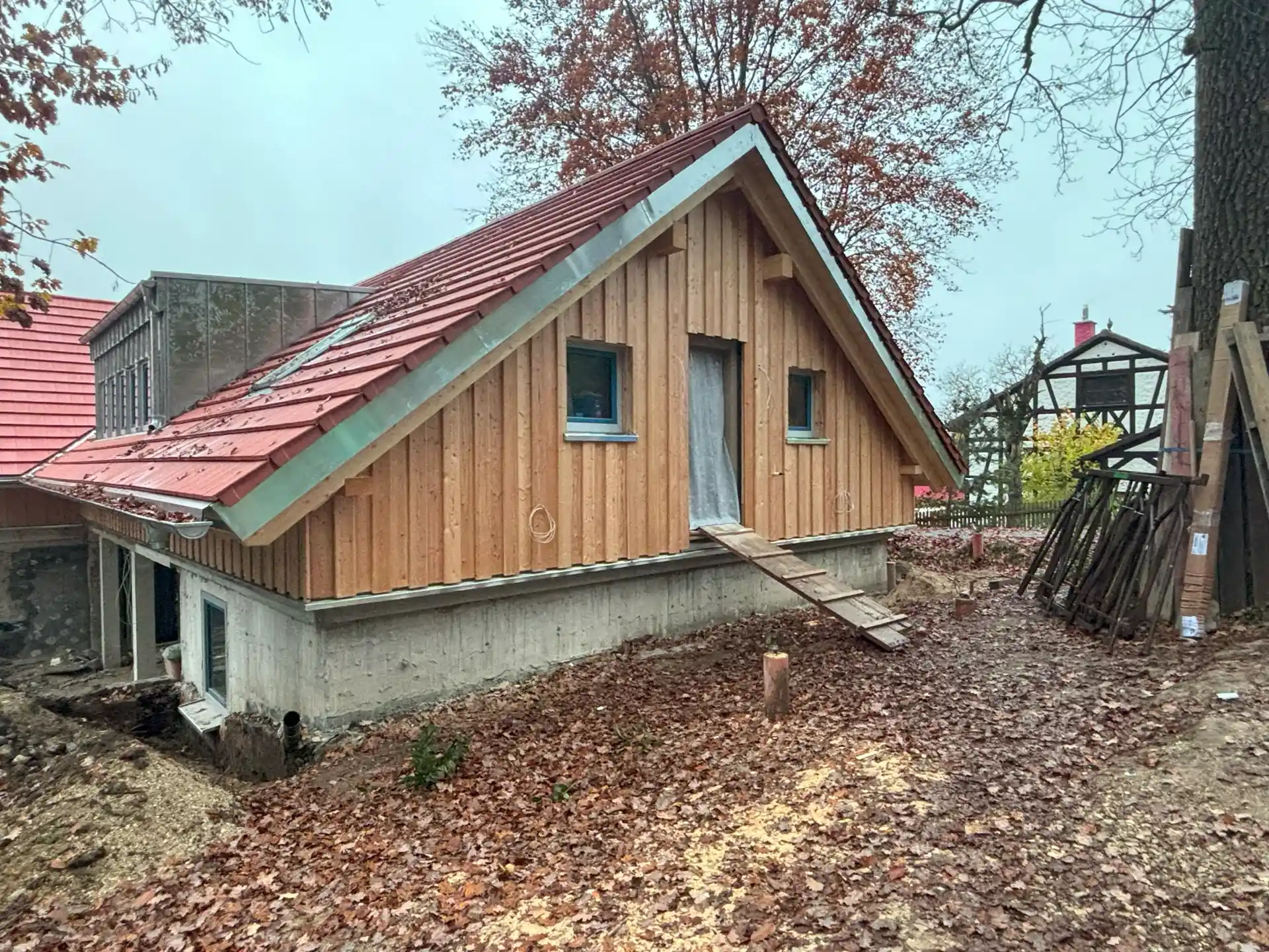 Neu saniertes Haus mit einer Holzfassade am Dachgeschoss und rotem Satteldach, im Vordergrund ist eine Erdfläche mit Herbstlaubt bedeckt.