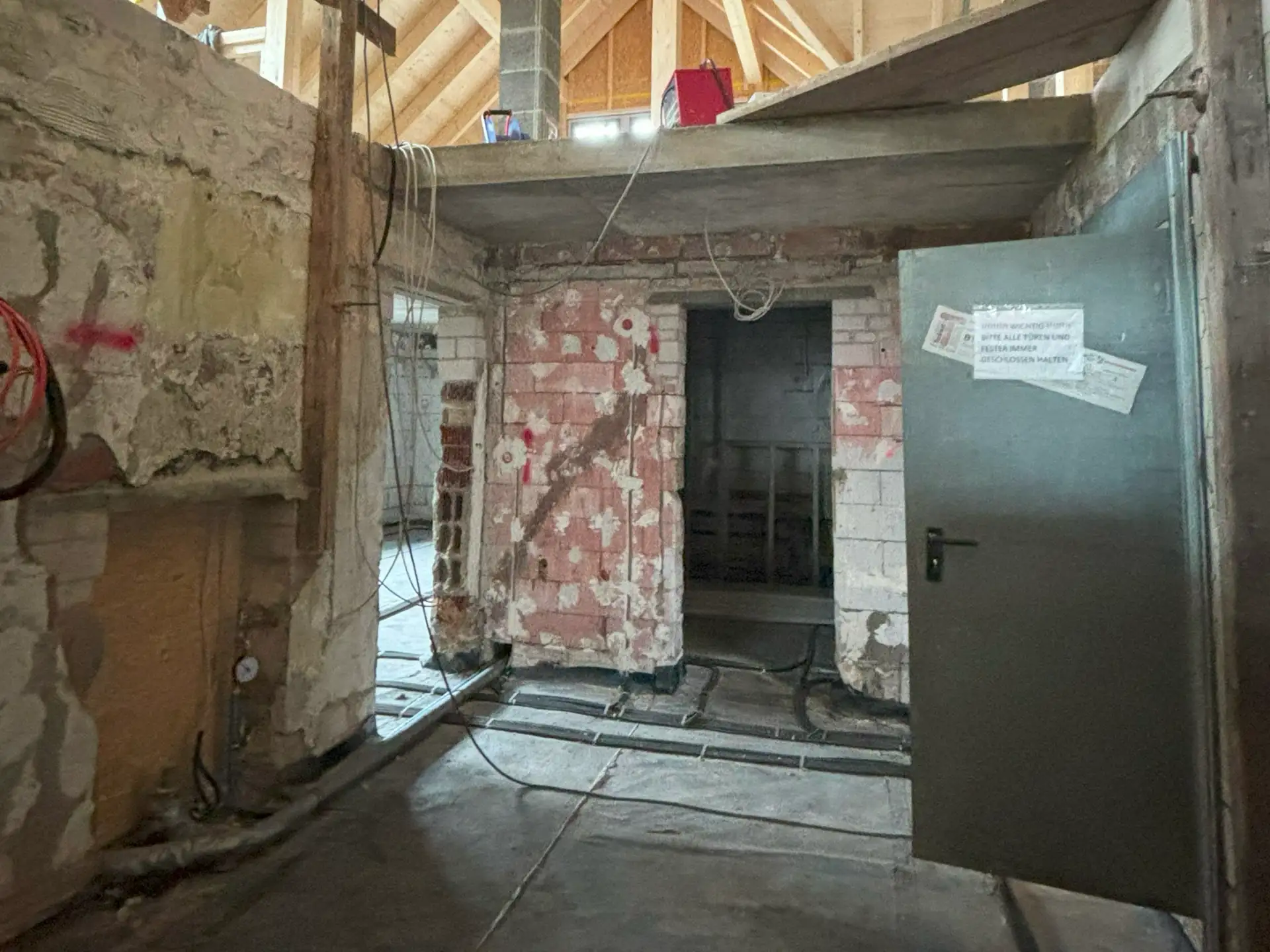 Rohbau eines Gebäudes nach Brandschaden während der Sanierung, mit freiliegenden Kabeln, unverputzten Wänden und einer offenen Tür mit angehängtem Zettel.