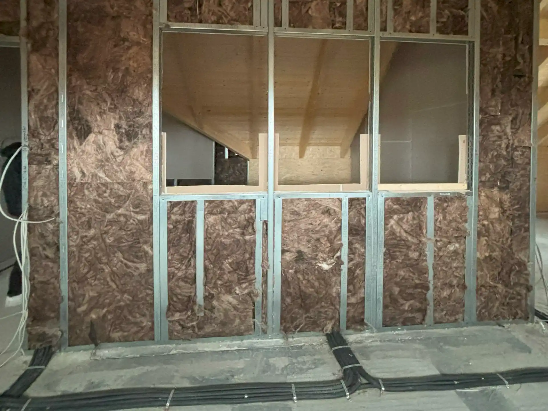 Innenwand mit Dämmstoff zwischen Metallständern, zwei rechteckige Fensteröffnungen, sichtbare Kabel auf dem Boden, Baustelle nach Brandschaden