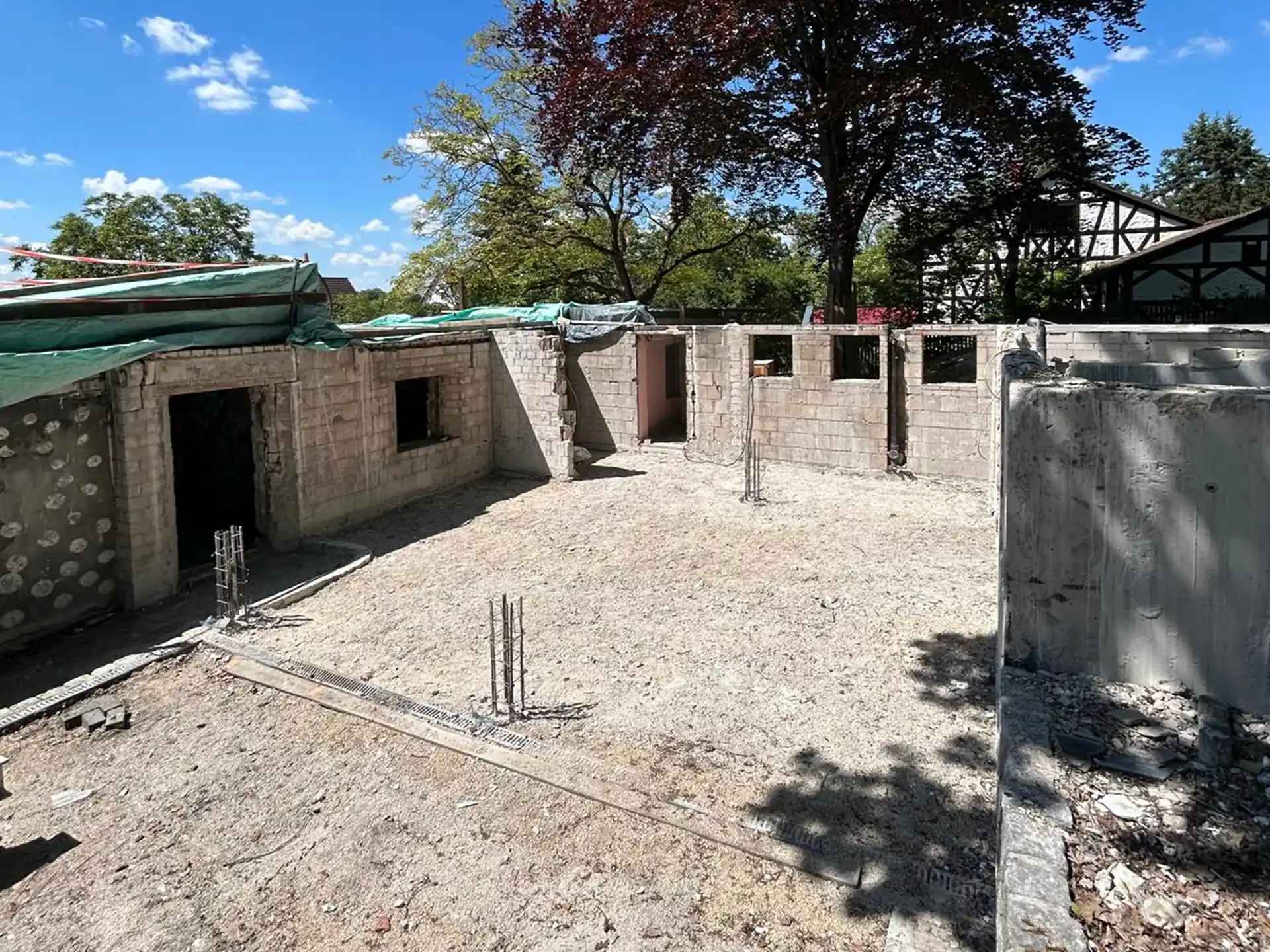 Rohbau mit Mauerwerk und Fensteröffnungen unter blauem Himmel, umgeben von Bäumen und Fachwerkhaus im Hintergrund.