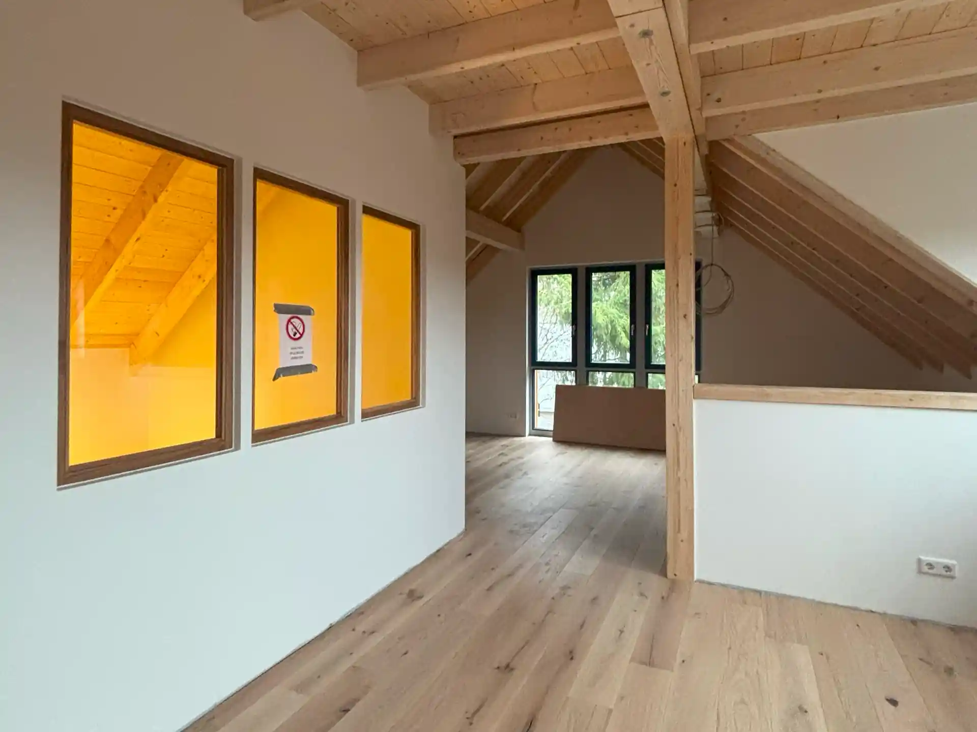 Heller Dachraum mit sichtbaren Holzbalken, drei gelb beleuchtete Fenster mit 'Rauchen verboten'-Schild, Holzboden und großer Fensterfront mit Blick ins Grüne.
