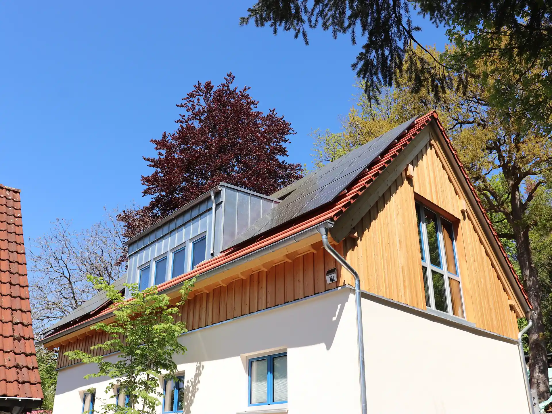Haus mit weißem Putz und Holzverkleidung im Dachgeschoss, Dach mit Solarpaneelen, umgeben von Bäumen unter blauem Himmel.