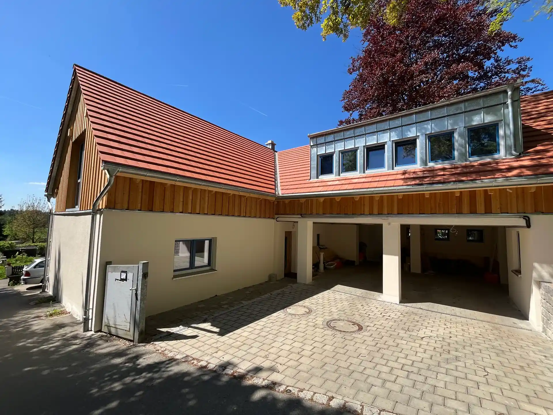 Haus mit neuem roten Ziegeldach, weißer Fassade und offener Garage, davor gepflasterte Einfahrt, im Hintergrund Baum mit rotem Laub.