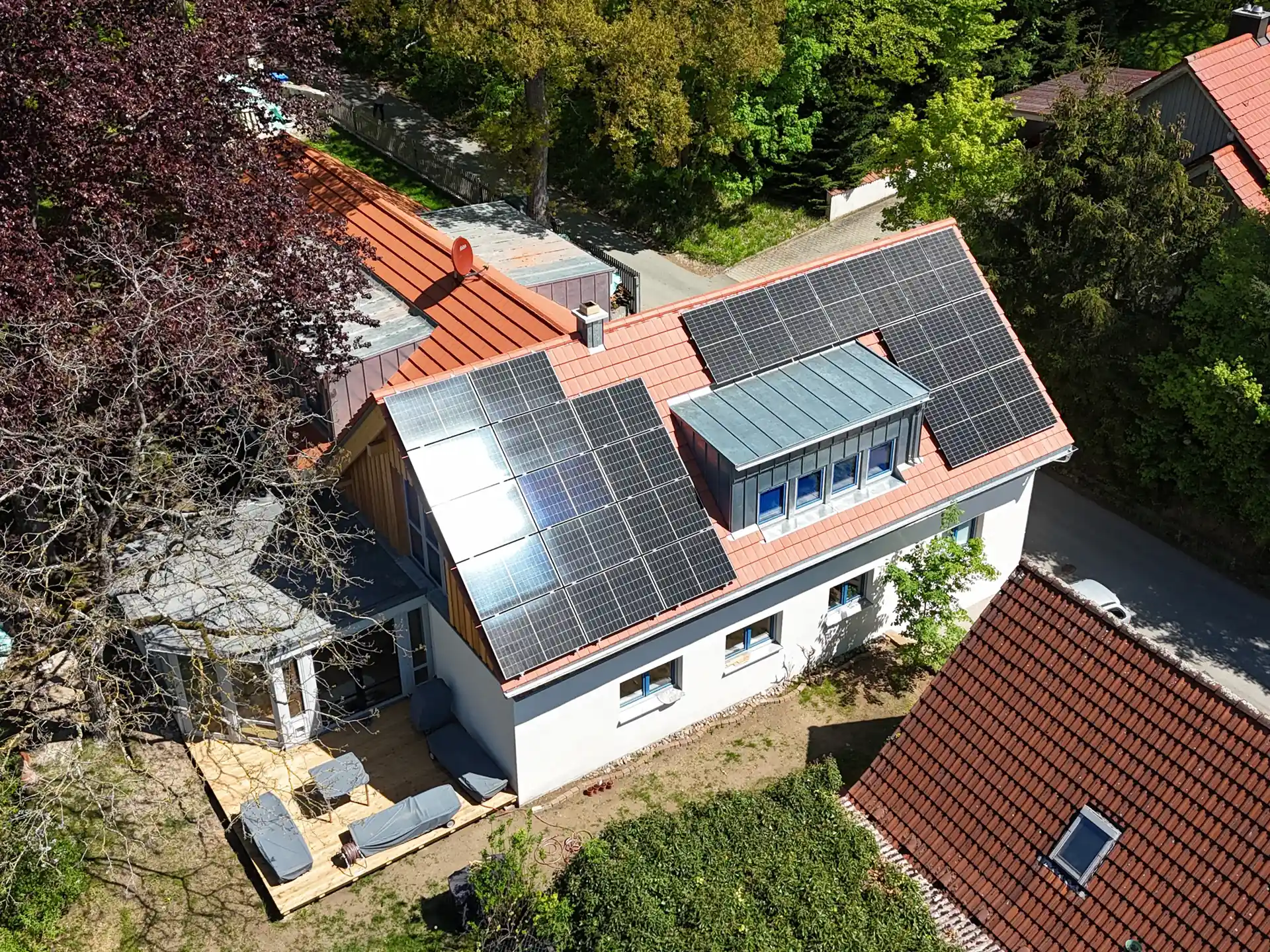 Luftaufnahme eines Hauses mit neuem roten Satteldach und mehreren schwarzen Solarpanelen, umgeben von Bäumen und Nachbargebäuden.