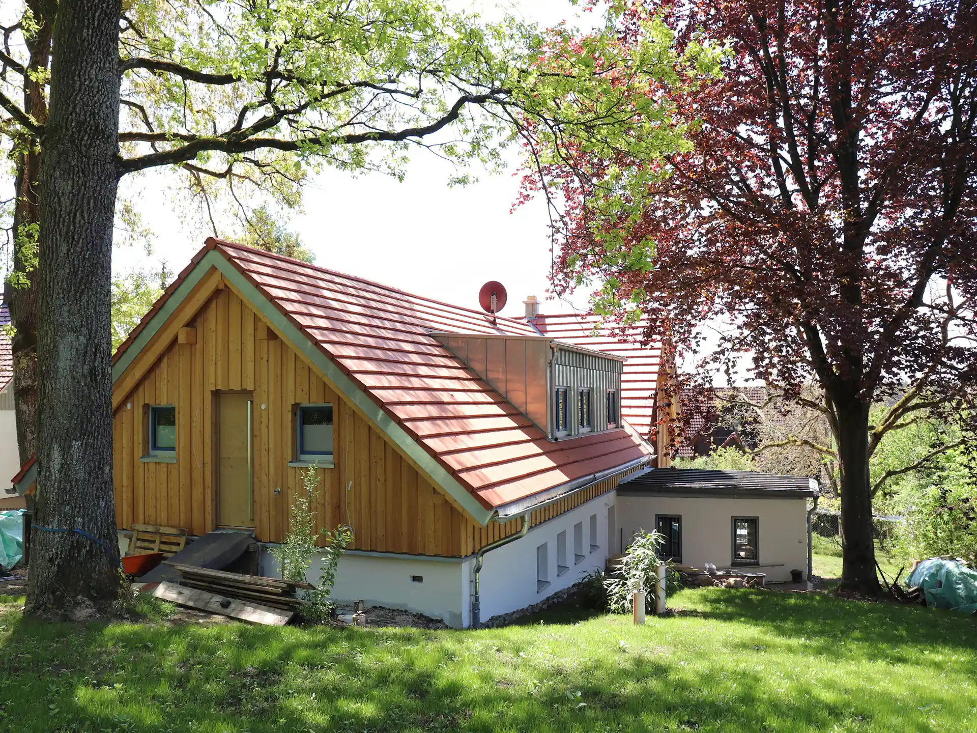 Neu saniertes Haus mit einer Holzfassade am Dachgeschoss und rotem Satteldach, im Vordergrund ist eine grüne Wiese. Das Haus ist von Bäumen umgeben.