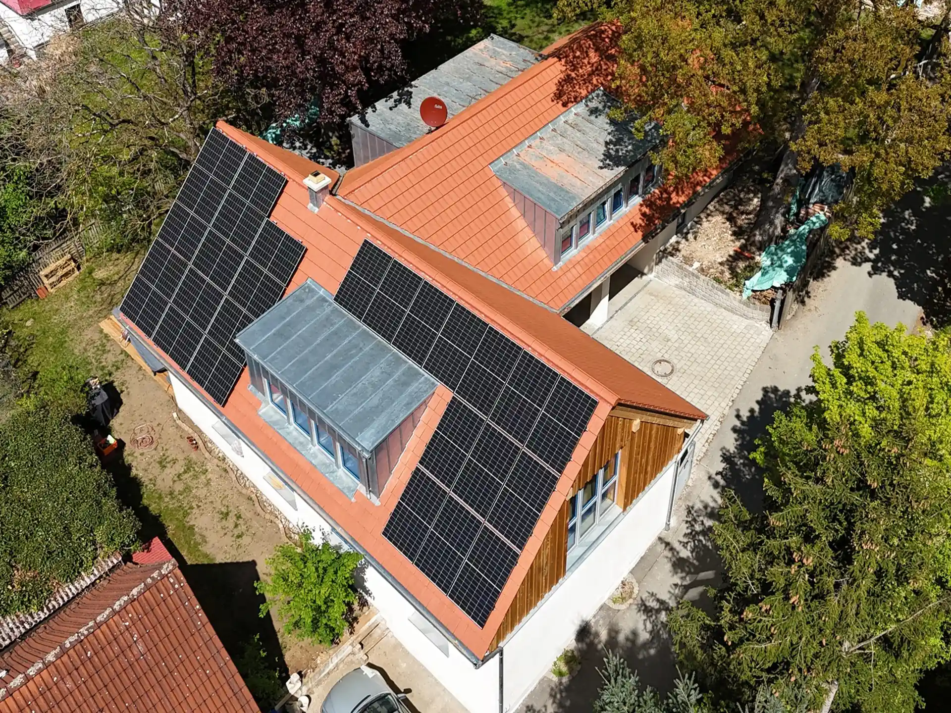 Luftaufnahme eines Hauses mit neuem roten Satteldach und mehreren schwarzen Solarpanelen, umgeben von Bäumen und Nachbargebäuden.