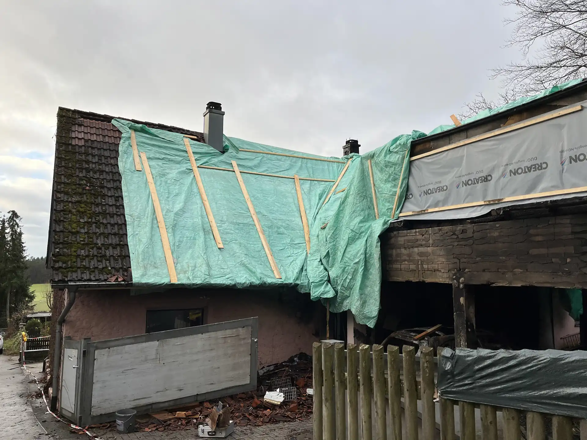 Zerstörtes Haus mit teilweise eingestürztem Dach, das mit grünem Planenmaterial und Holzlatten abgedeckt ist.