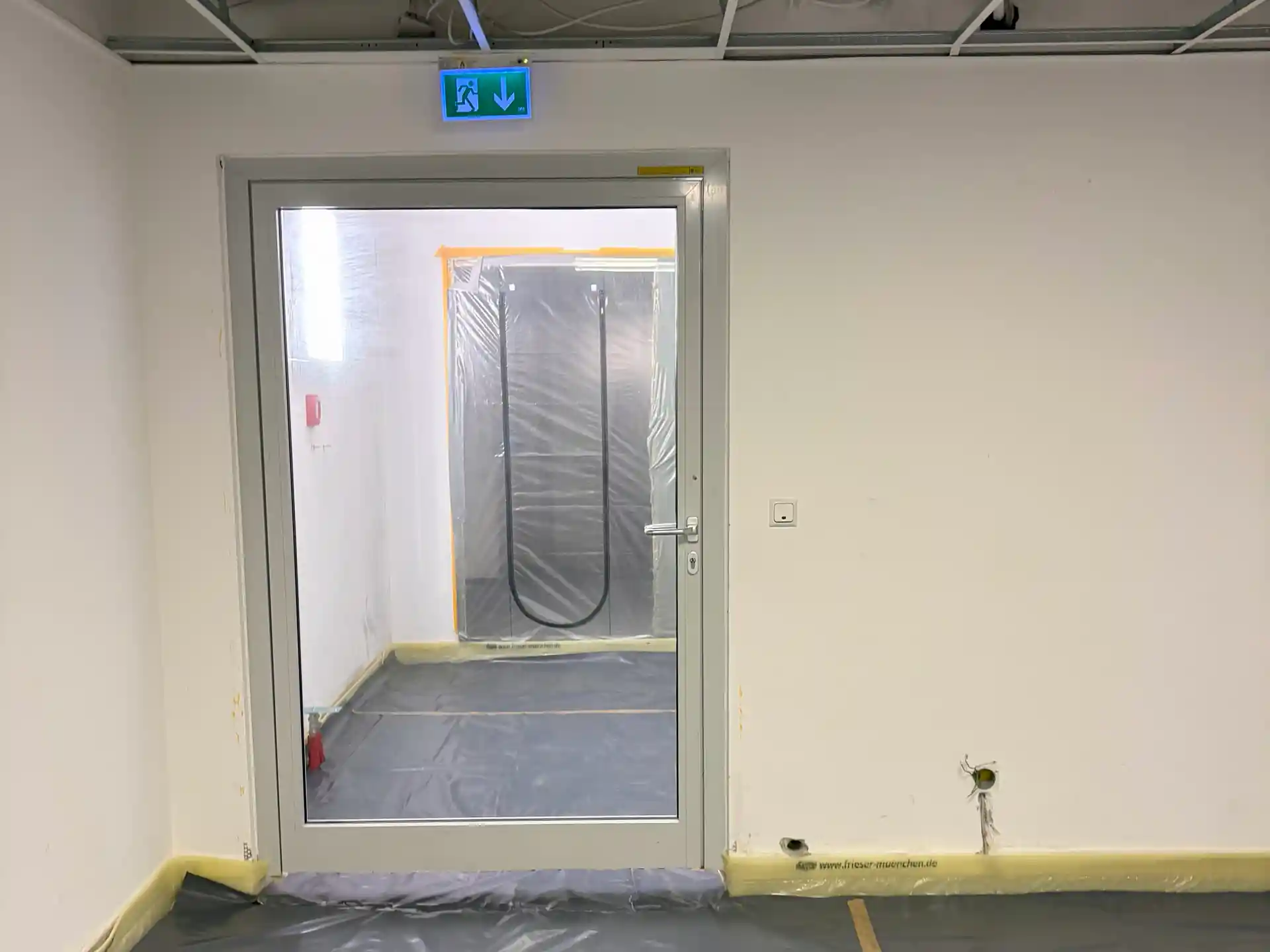 Raum mit weiß gestrichenen Wänden und Tür mit Glasfenster, dahinter ein weiterer Raum mit Edelstahltür in Schutzfolie über der Tür und auf dem Boden, Notausgangsschild über der Glastür.