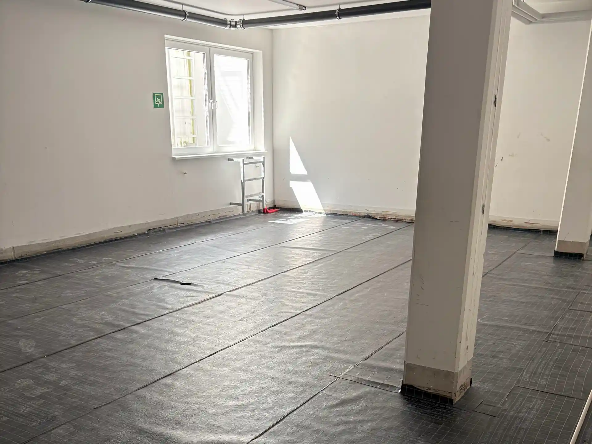 Leerer Raum mit weiß gestrichenen Wänden, zwei weißen Stützsäulen und schwarzer Schutzfolie auf dem Boden, Fenster mit Gitter und Notausgangsschild an der Wand.