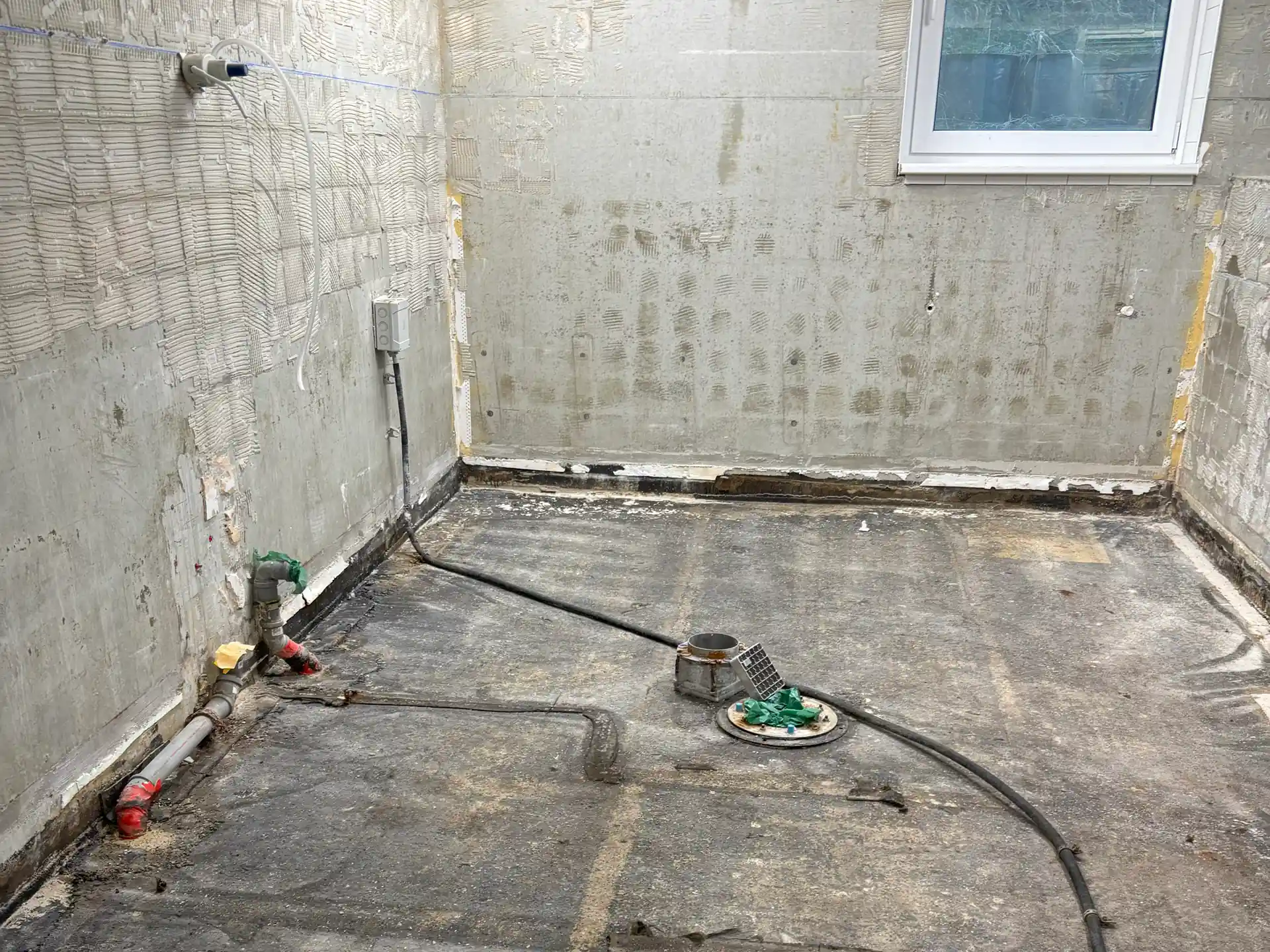 Leerer Raum mit freigelegtem Estrich, sichtbaren Rohrleitungen und Abfluss in der Mitte, Wände mit Putzresten und Fenster rechts oben.