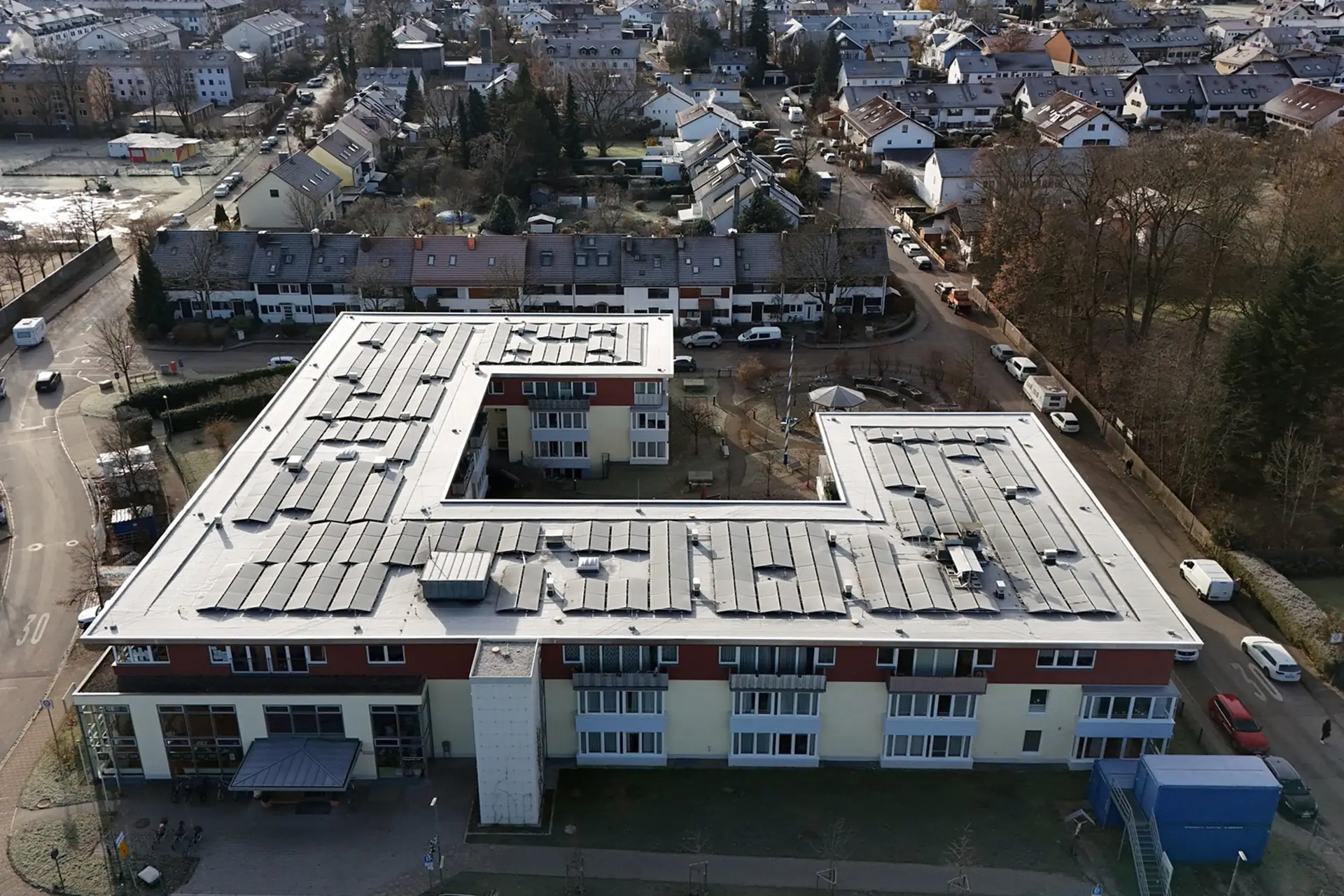 Luftaufnahme eines großen, flachen Wohngebäudes, umgeben von weiteren Wohnhäusern und Bäumen.
