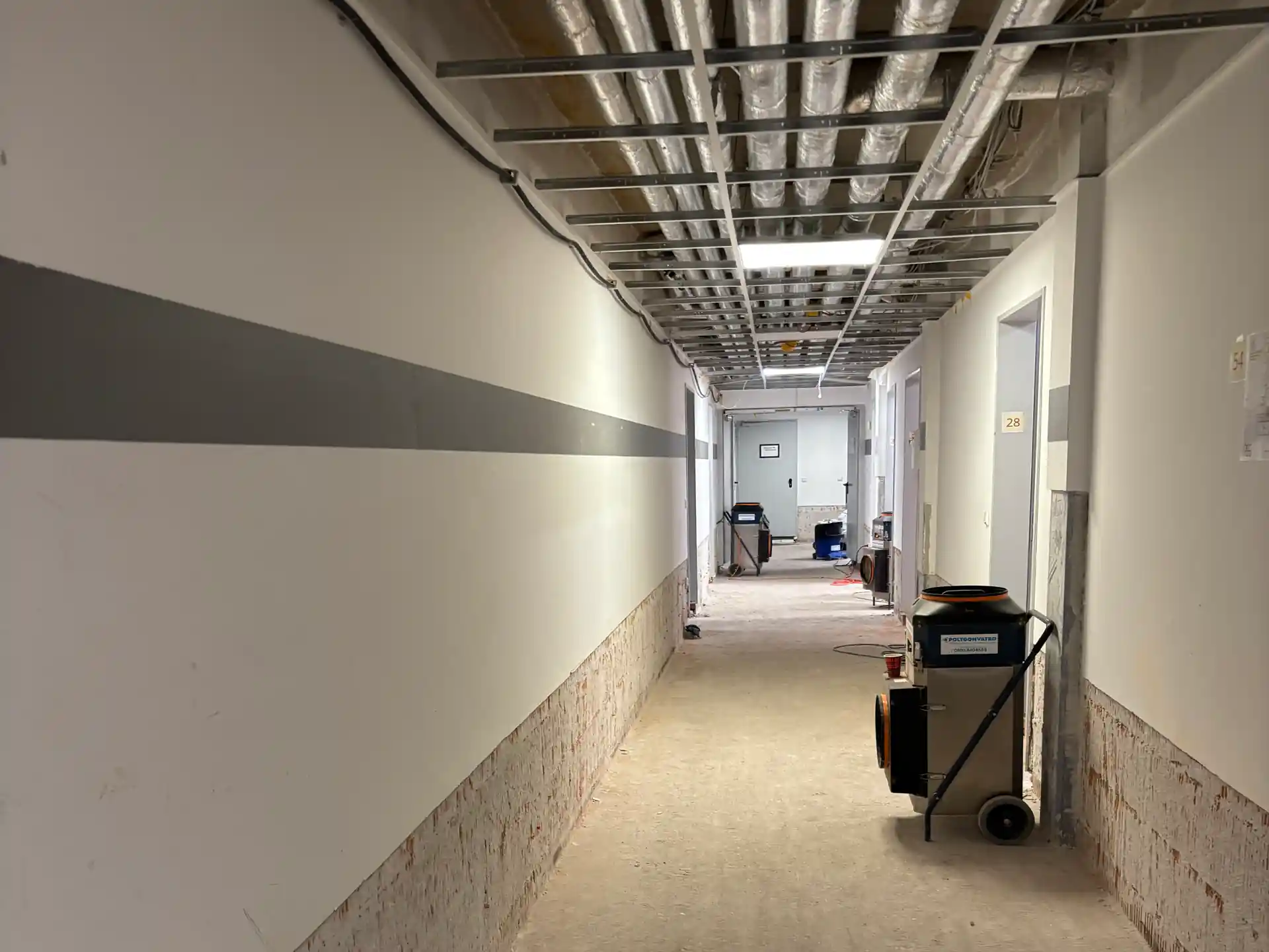 Flur mit abgehängter Decke und sichtbaren Leitungen, Wände unten freigelegt, Baustellengeräte auf dem Boden, Türen rechts mit Nummer 28.