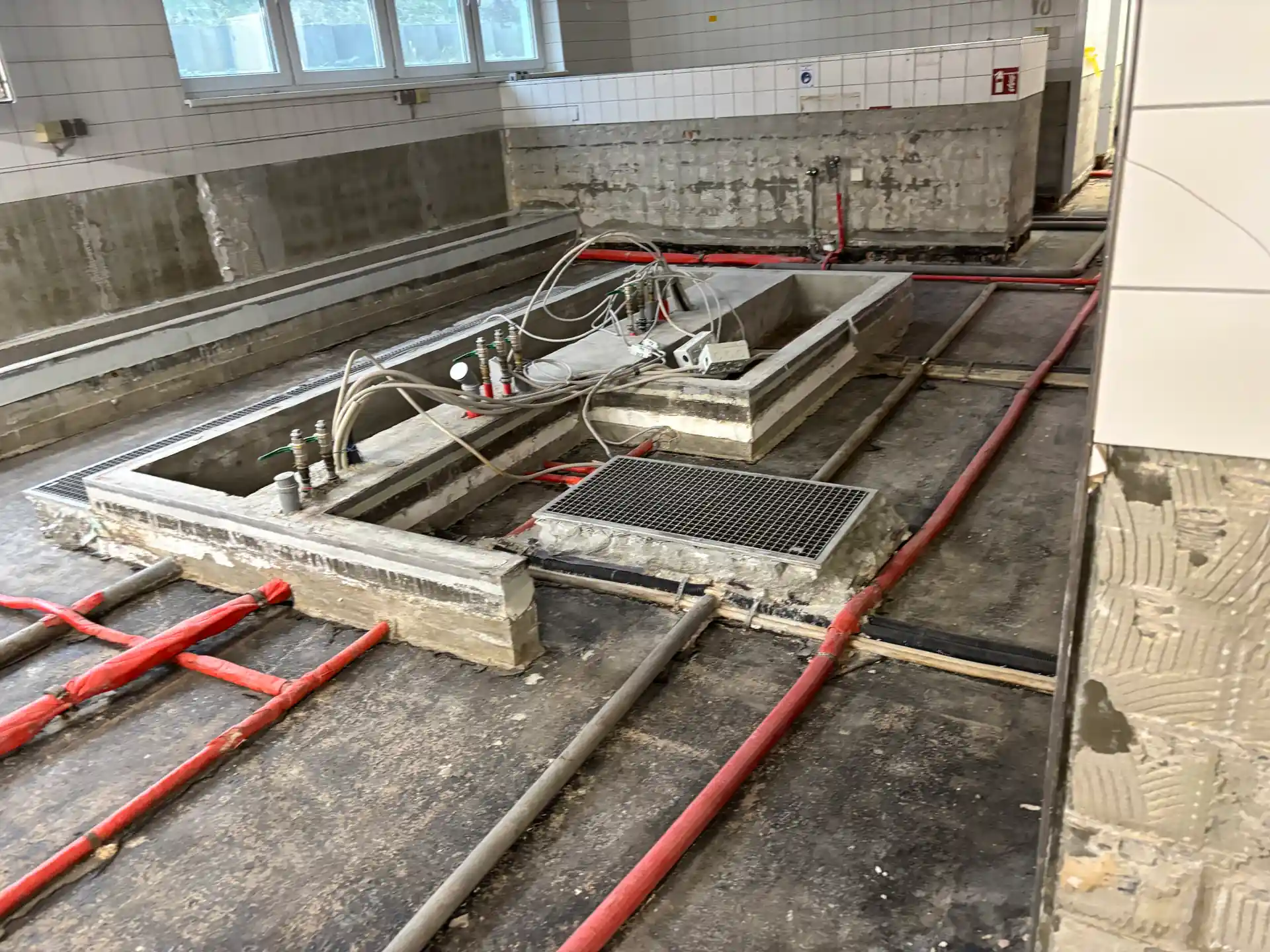 Boden mit freigelegten Leitungen und Rohren in einem Raum mit weißen Fliesen und Fenstern, Sanierung nach Elementarschaden.