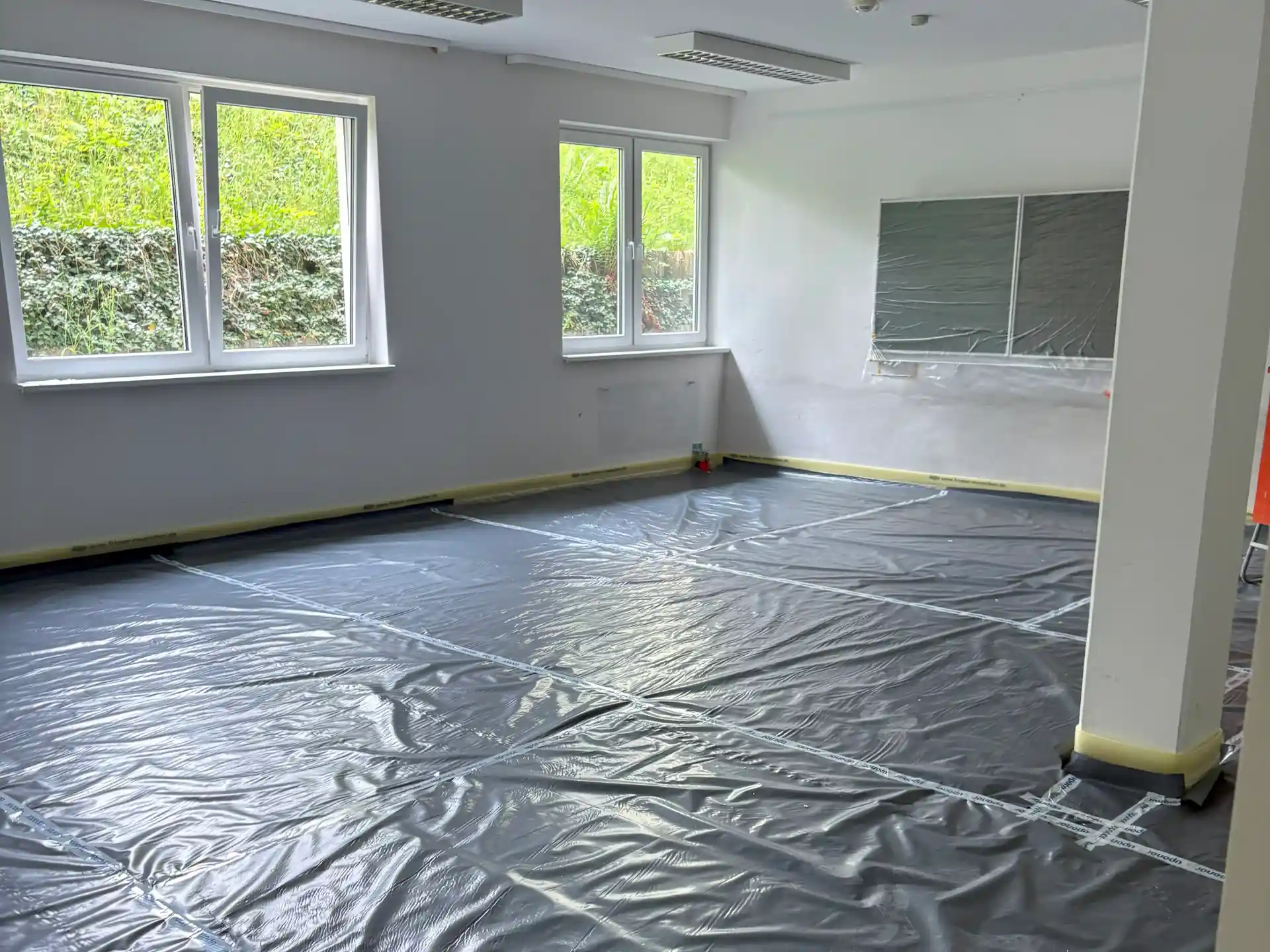 Leerer Raum mit weiß gestrichenen Wänden, zwei weißen Stützsäulen und schwarzer Schutzfolie auf dem Boden, Fenster mit Blick ins Grüne.