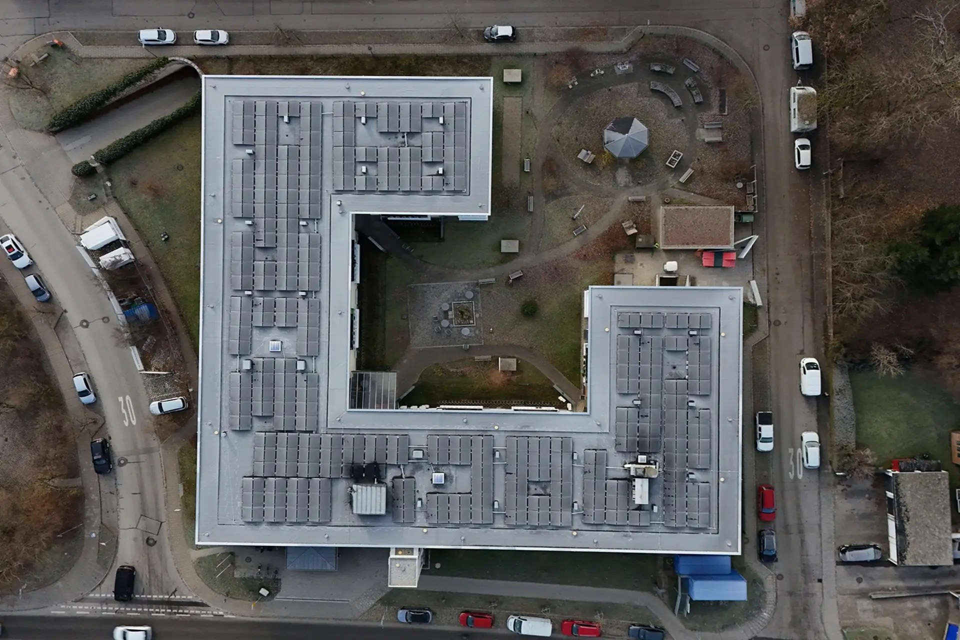 Luftaufnahme eines großen, flachen Wohngebäudes, umgeben von Straßenzügen, parkenden Autos und Bäumen.