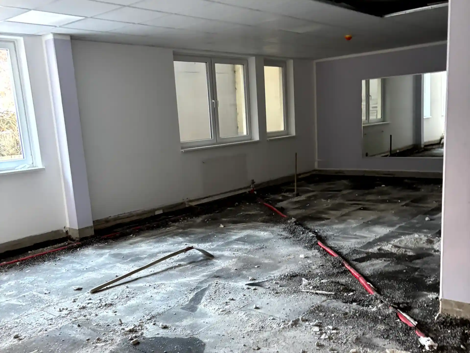 Leerer Raum mit beschädigtem Boden, freiliegenden roten Rohren und weißen Wänden, drei Fenster an der linken Wand.