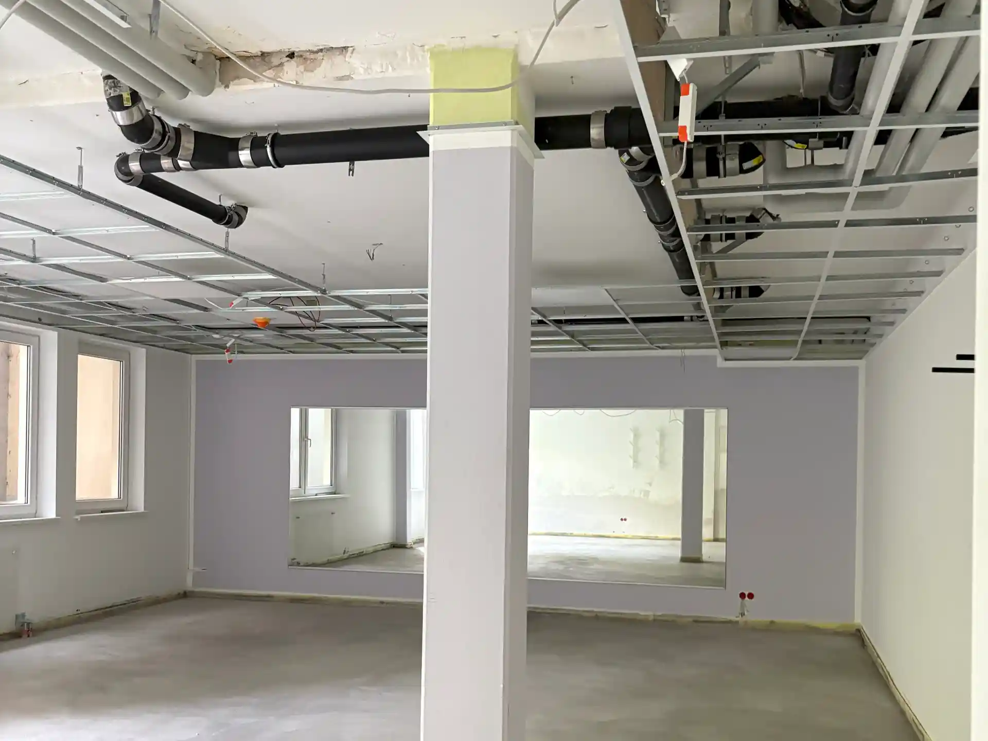 Leerer Raum mit weiß gestrichener Säule in der Mitte, sichtbaren Rohrleitungen an der Decke und großem rechteckigem Fenster in der Wand.
