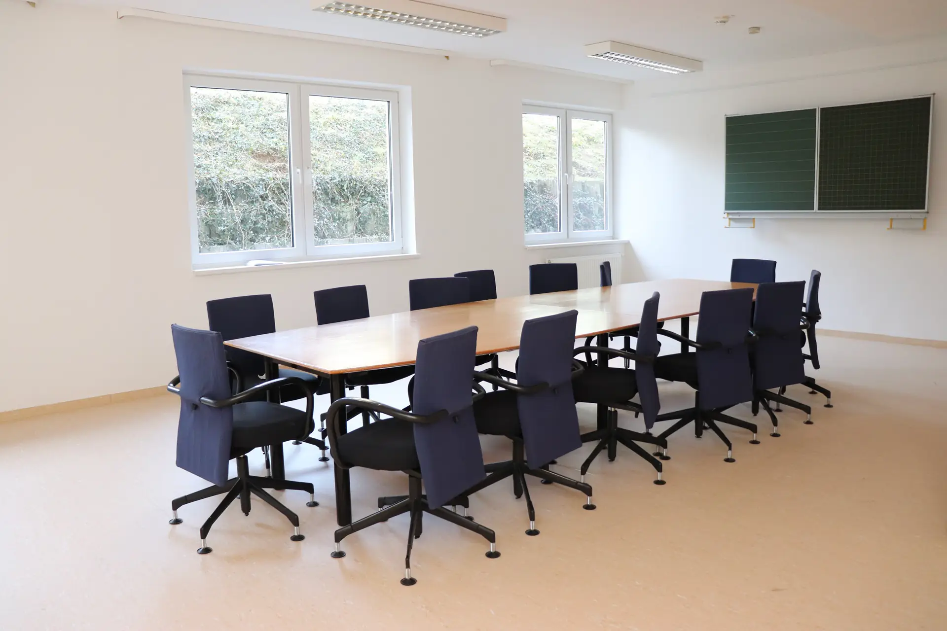 Großer rechteckiger Konferenztisch mit mehreren schwarzen Bürostühlen in einem hellen Raum mit zwei Fenstern und zwei grünen Tafeln an der Wand.