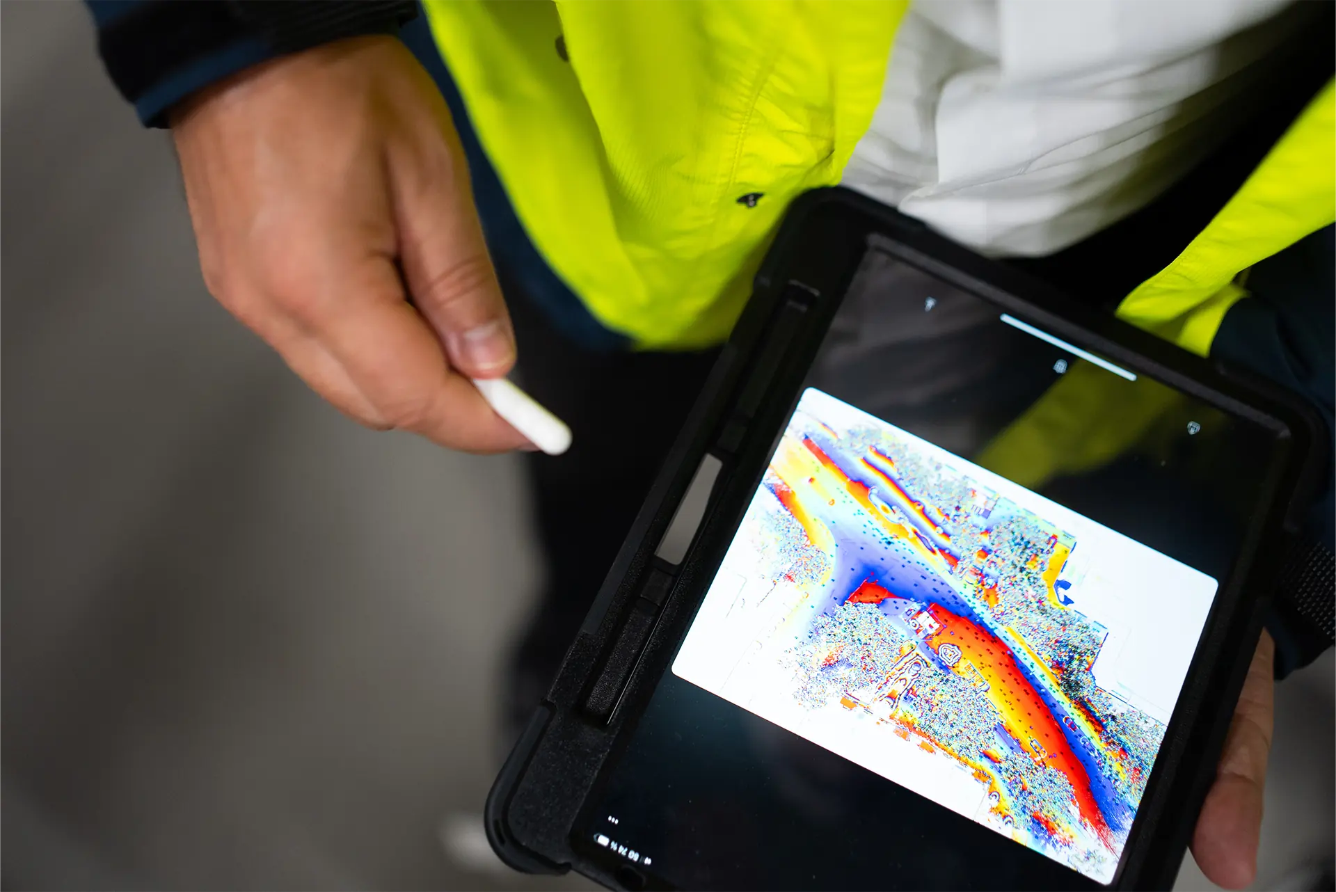 Eine Person in gelber Sicherheitsjacke hält in der linken Hand ein Tablet, auf dem Daten visualisiert dargestellt werden. In der rechten Hand hält die Person einen Stylus.