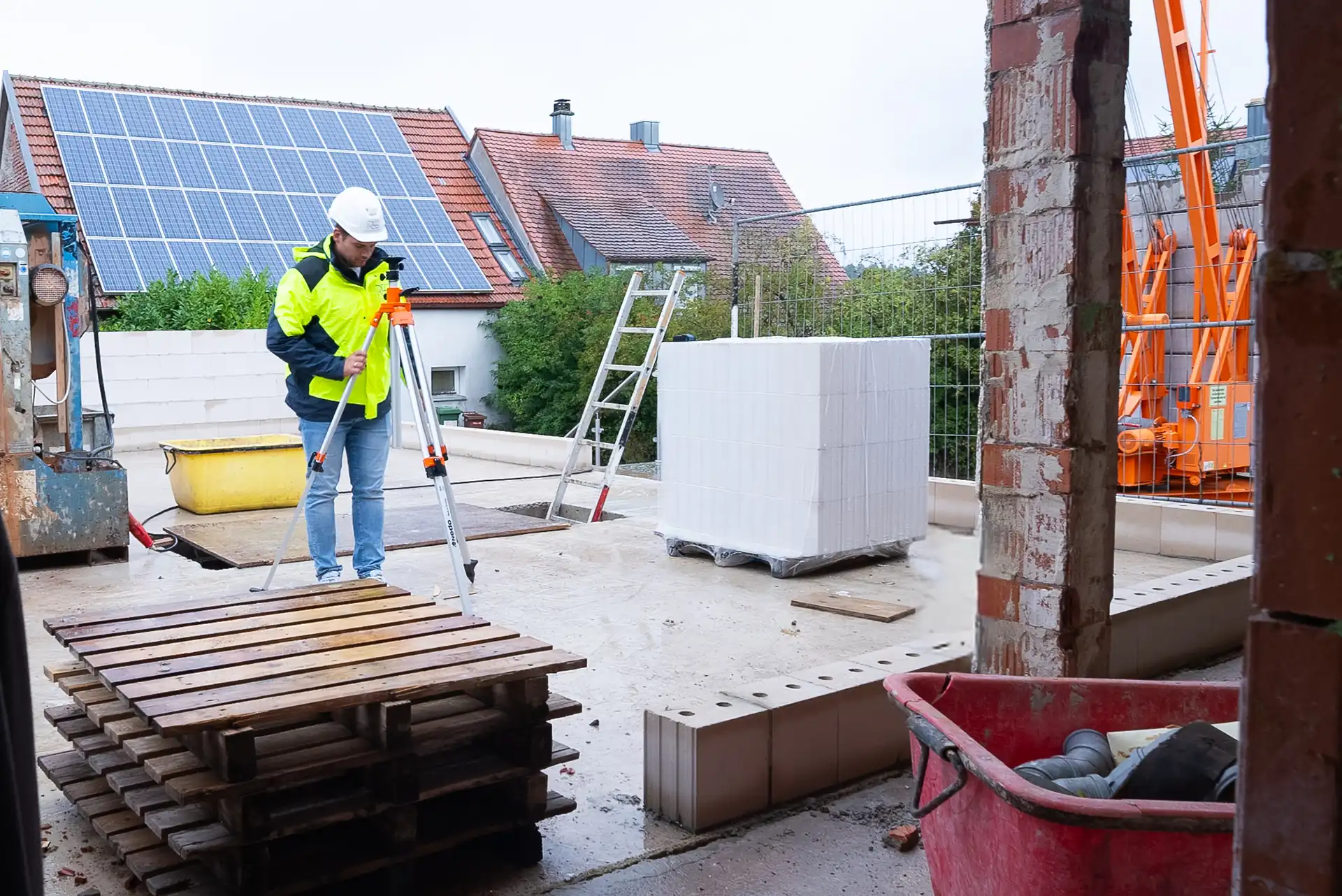 Blick durch einen offenen Türrahmen auf eine Baustelle. Ein Bauarbeiter in gelber Sicherheitsjacke und Helm steht auf einem Betonboden. Im Hintergrund sind Solarpanels und Baugeräte sichtbar.