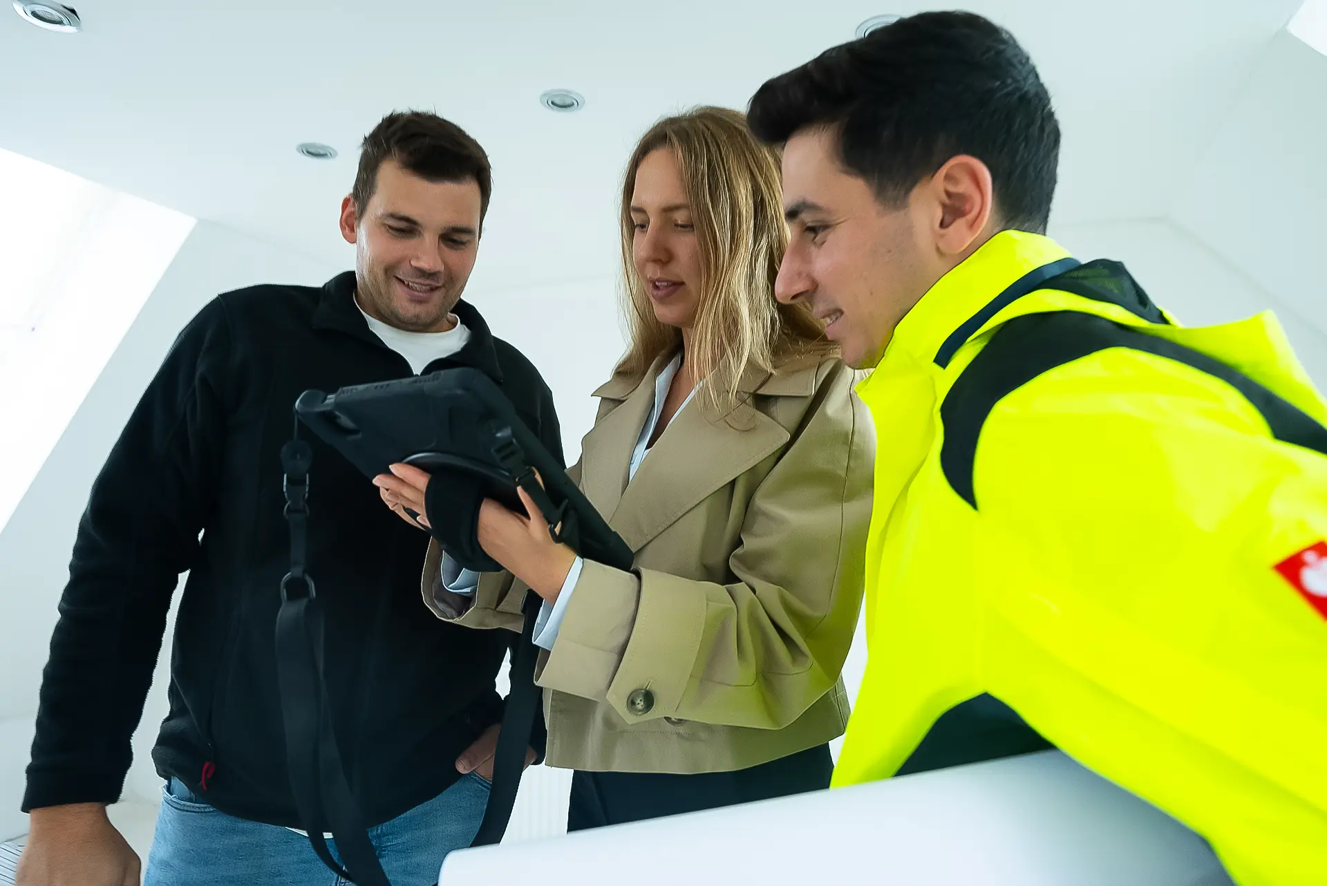 Drei Personen stehen zusammen, eine hält ein Tablet, eine trägt eine schwarze Jacke, eine eine beige Jacke, die dritte eine gelbe Sicherheitsjacke und hält eine Planrolle unter dem linken Arm.