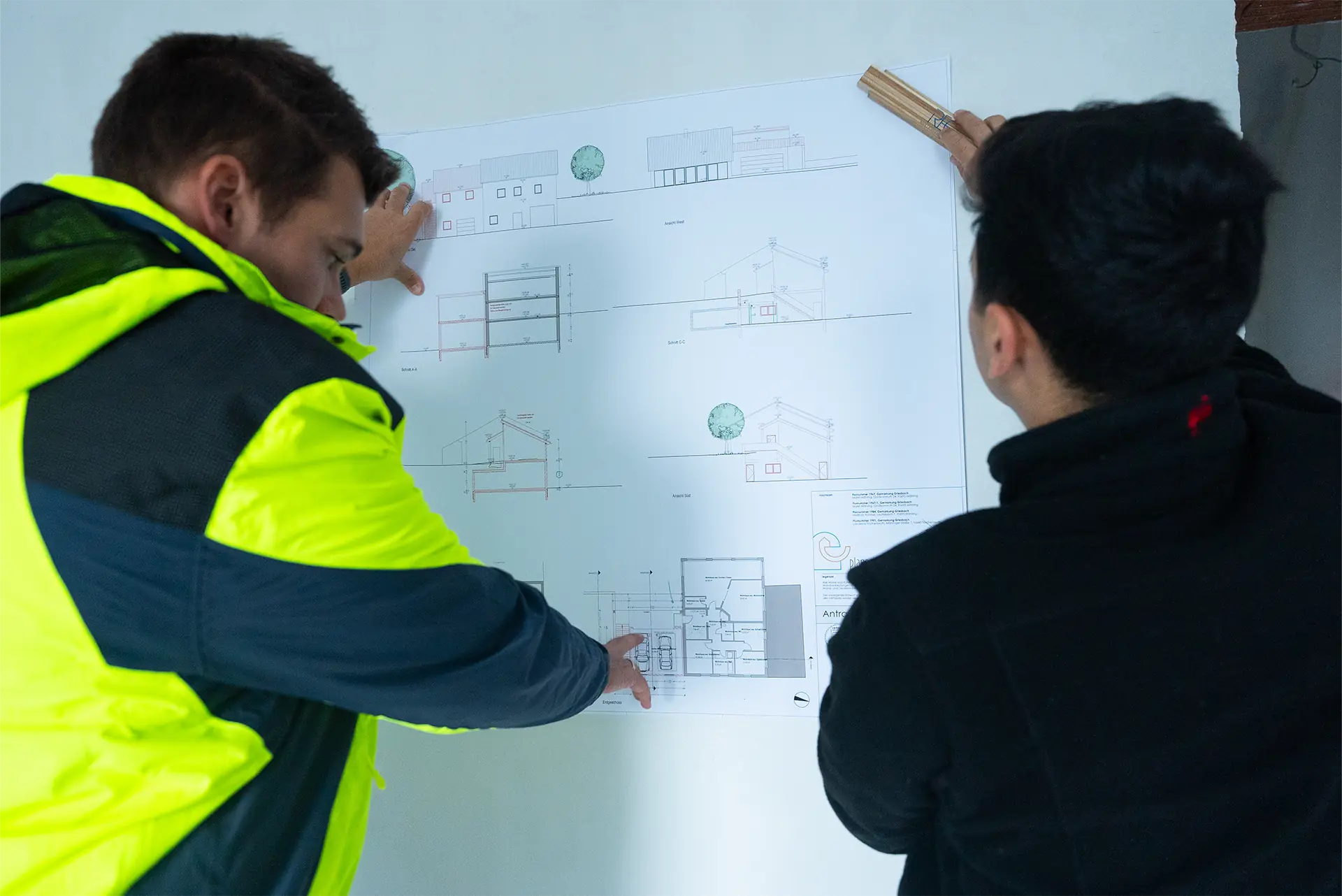 Zwei Männer betrachten einen Bauplan an einer weißen Wand, einer zeigt mit dem Finger auf den Plan, der andere hält ein Lineal.