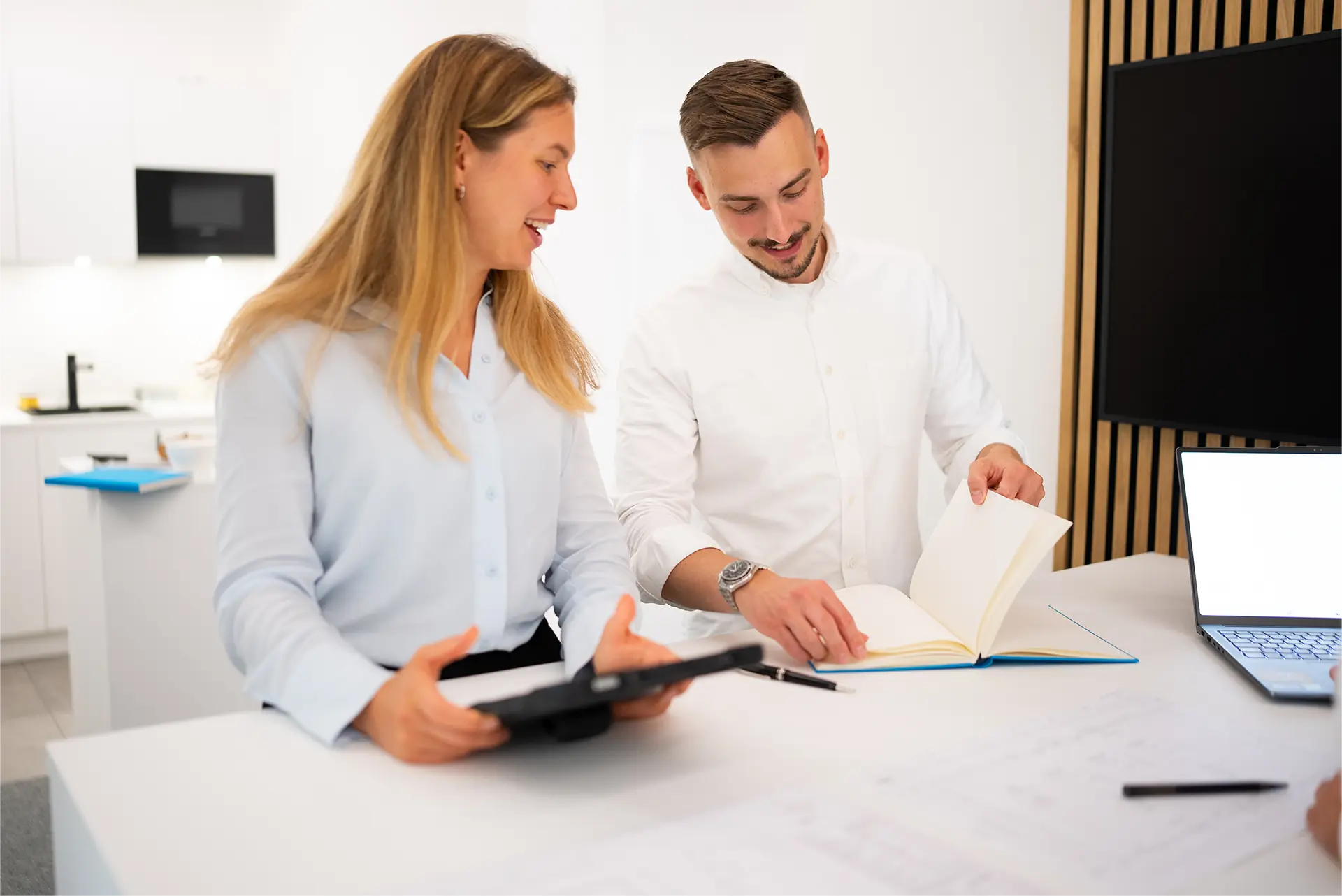 Zwei Personen stehen an einem weißen Tisch, eine hält ein Tablet, die andere blättert in einem Buch, im Hintergrund ein Bildschirm und eine weiße Wand.