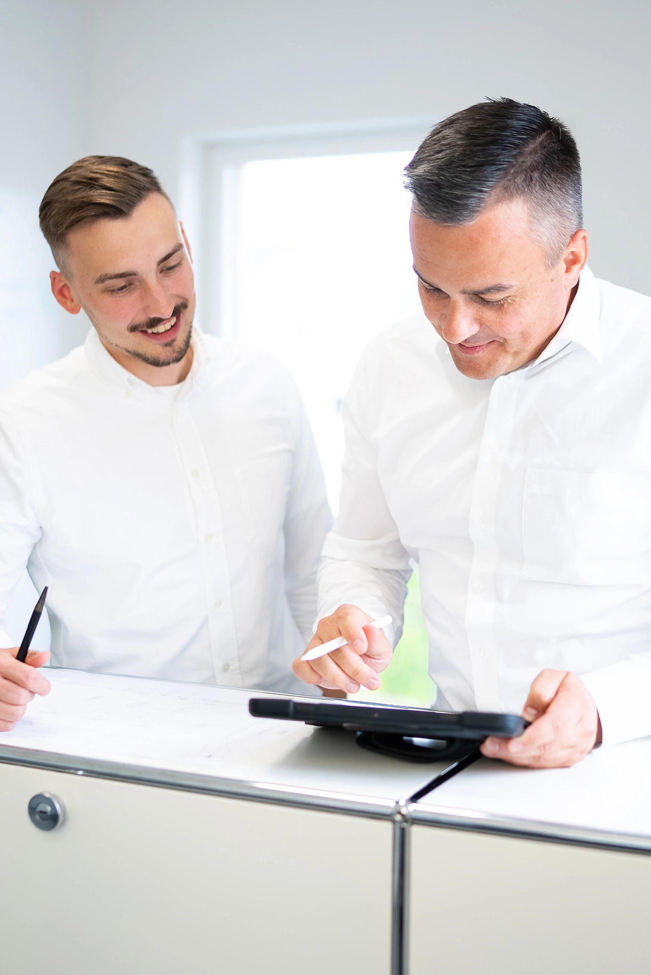 Zwei Männer in weißen Hemden stehen an einem Schreibtisch, einer hält einen Stift, der andere ein Tablet und einen Stylus.