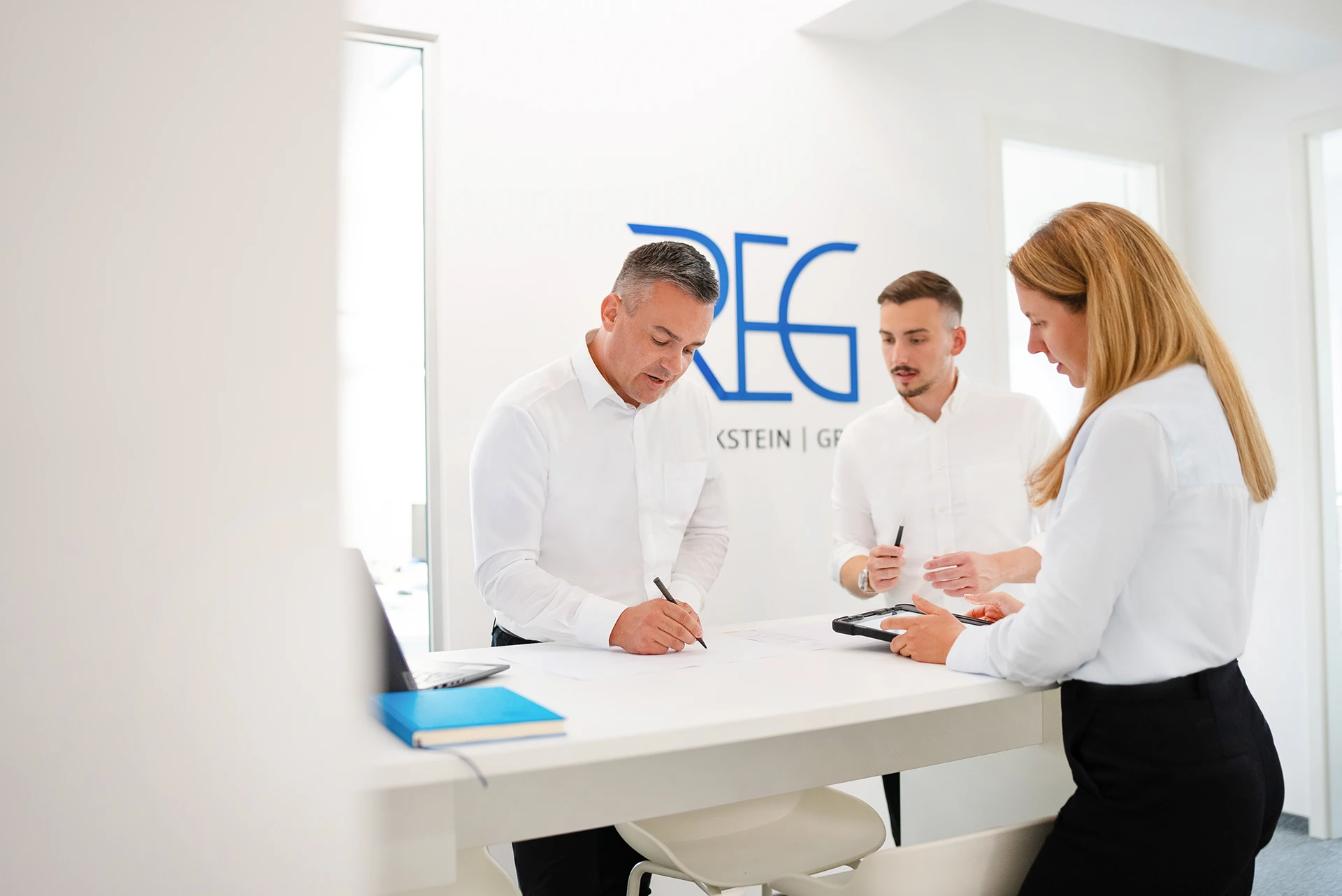 Drei Personen in weißem Hemd und Bluse stehen an einem weißen Tresen, im Hintergrund ein Logo mit blauen Buchstaben "REG".