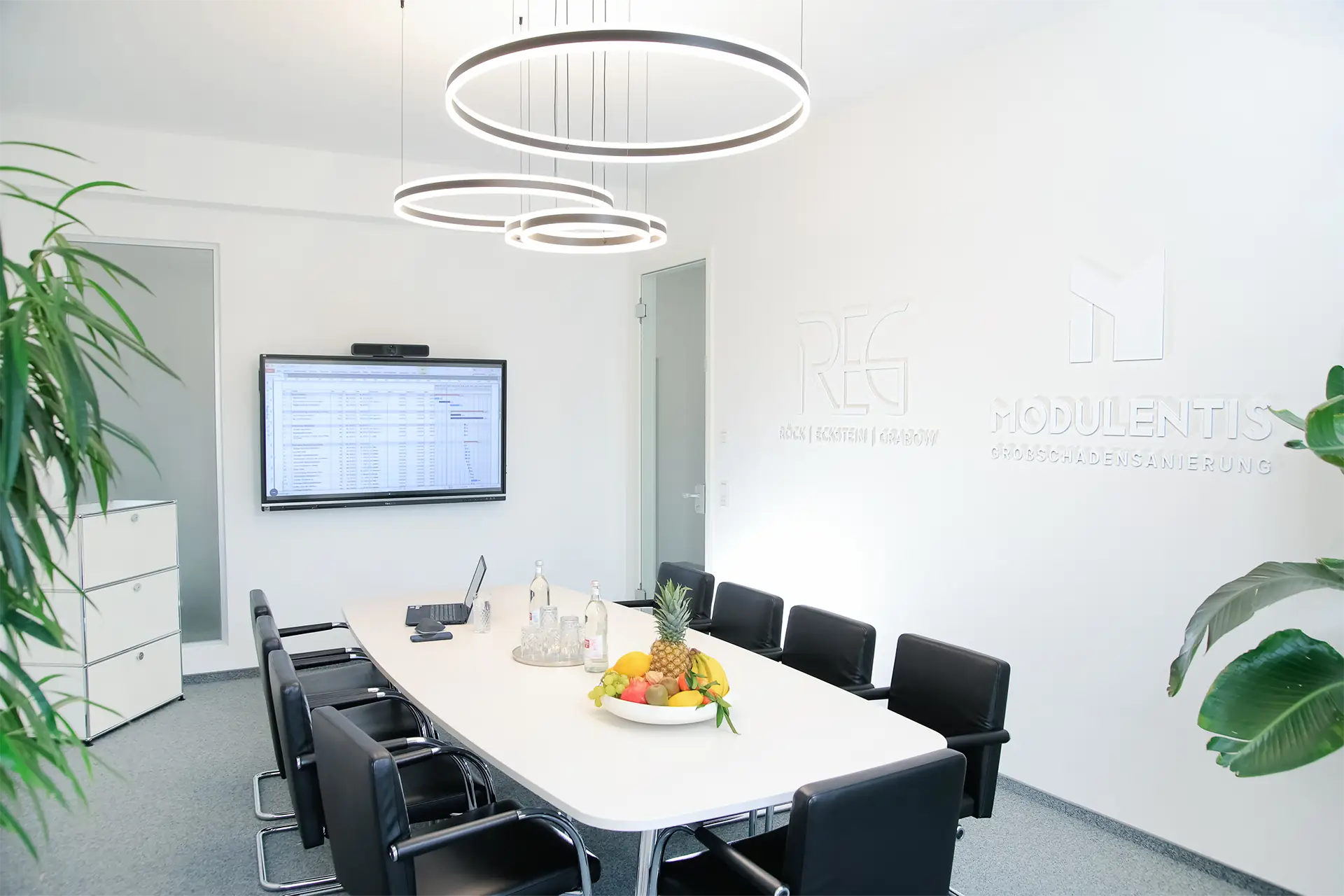 Moderner Besprechungsraum mit langem weißen Tisch, schwarzen Stühlen, Obstschale in der Mitte und zwei runden Deckenleuchten. An der hinteren Wand hängt ein Monitor.