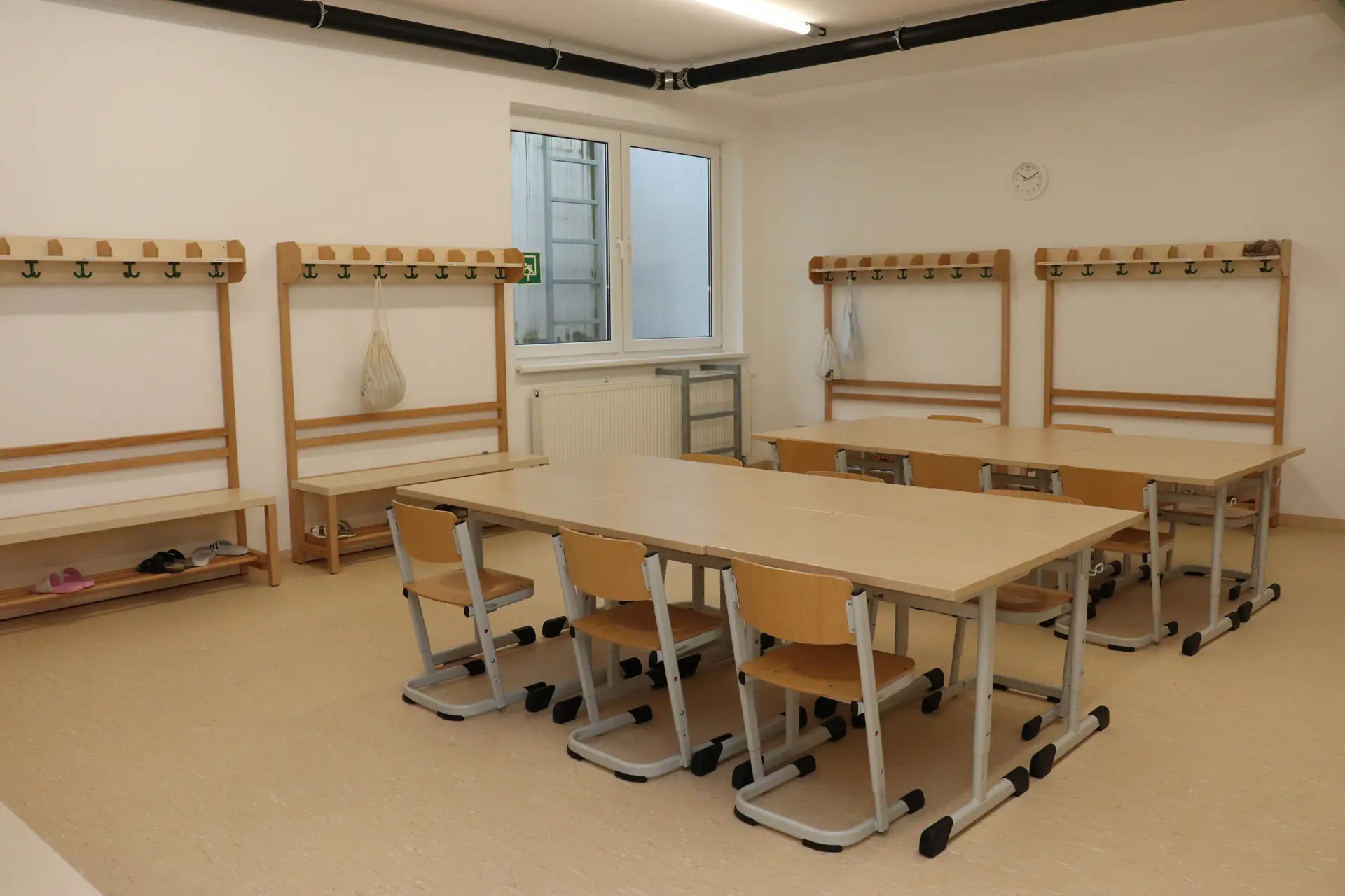 Leerer Umkleideraum mit mehreren Holzgarderoben, einem Fenster und zwei Tischeb mit jeweils sechs Stühlen in der Mitte.