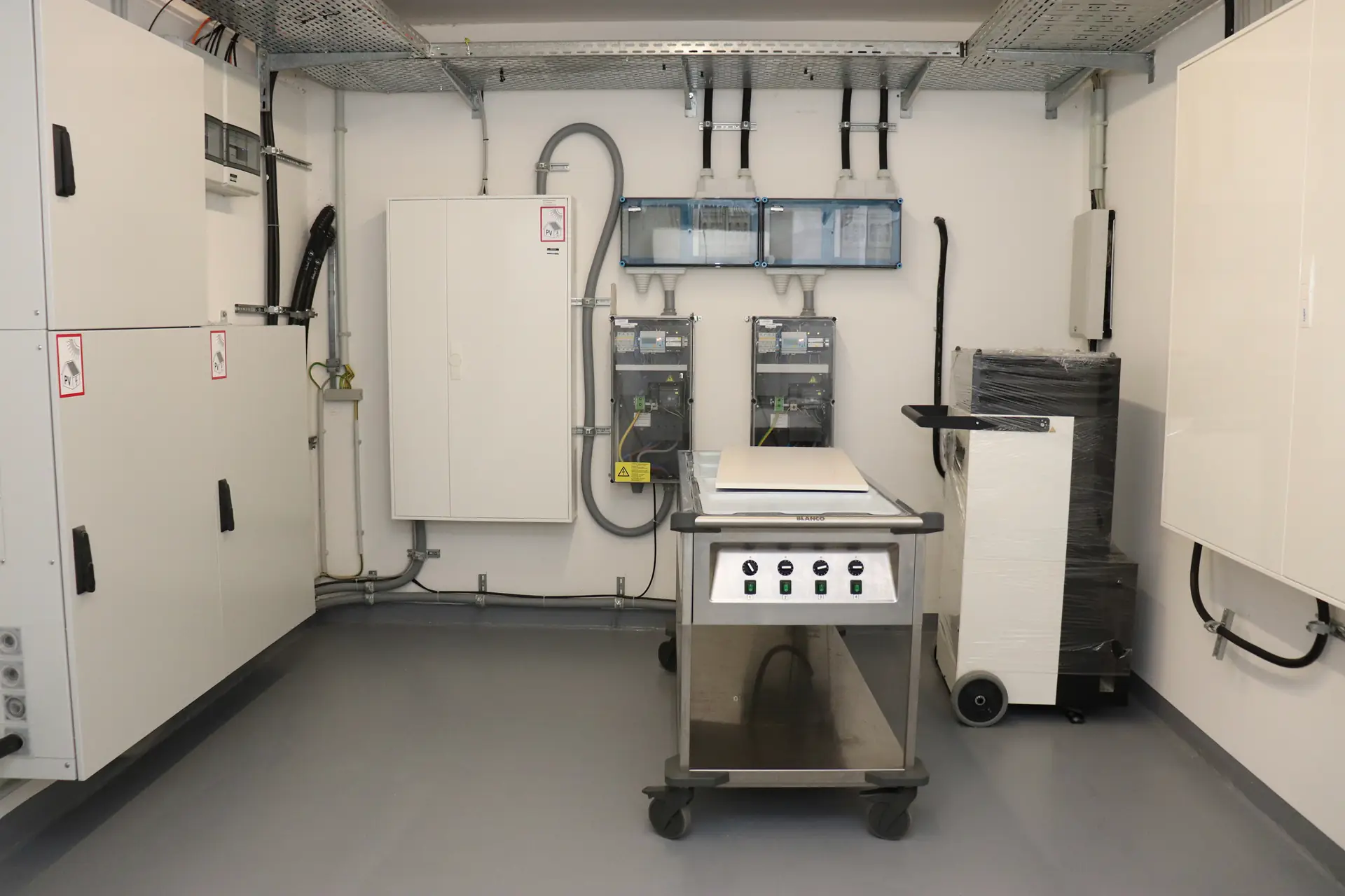 Technikraum mit mehreren weißen Schaltschränken an den Wänden und einem fahrbaren Metalltisch mit Bedienfeld in der Mitte.