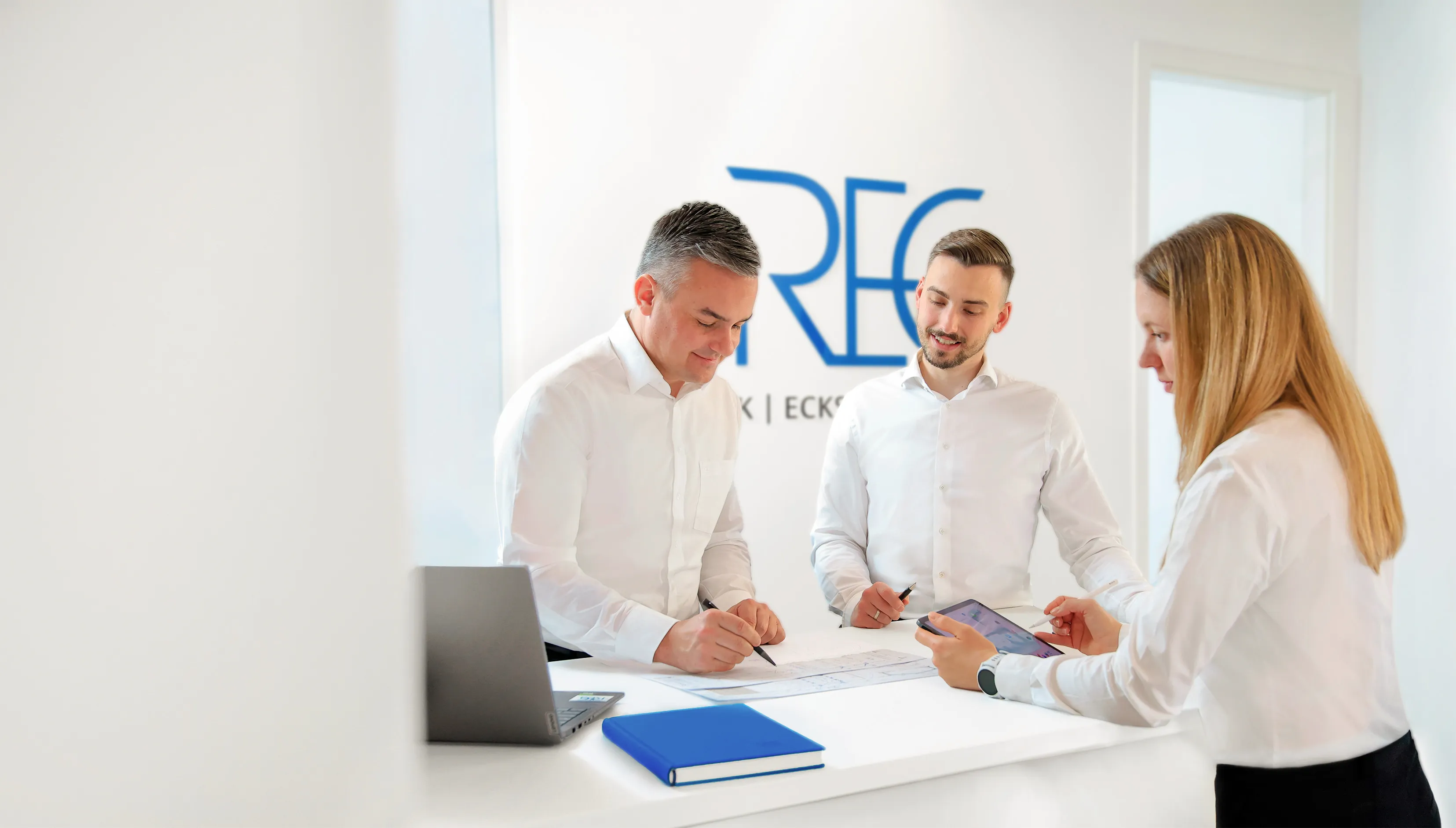Drei Mitarbeiter eines Ingenieurbüros besprechen gemeinsam Baupläne an einem hellen Schreibtisch in einem modernen Büro, im Hintergrund das REG-Ingenieure-Logo.