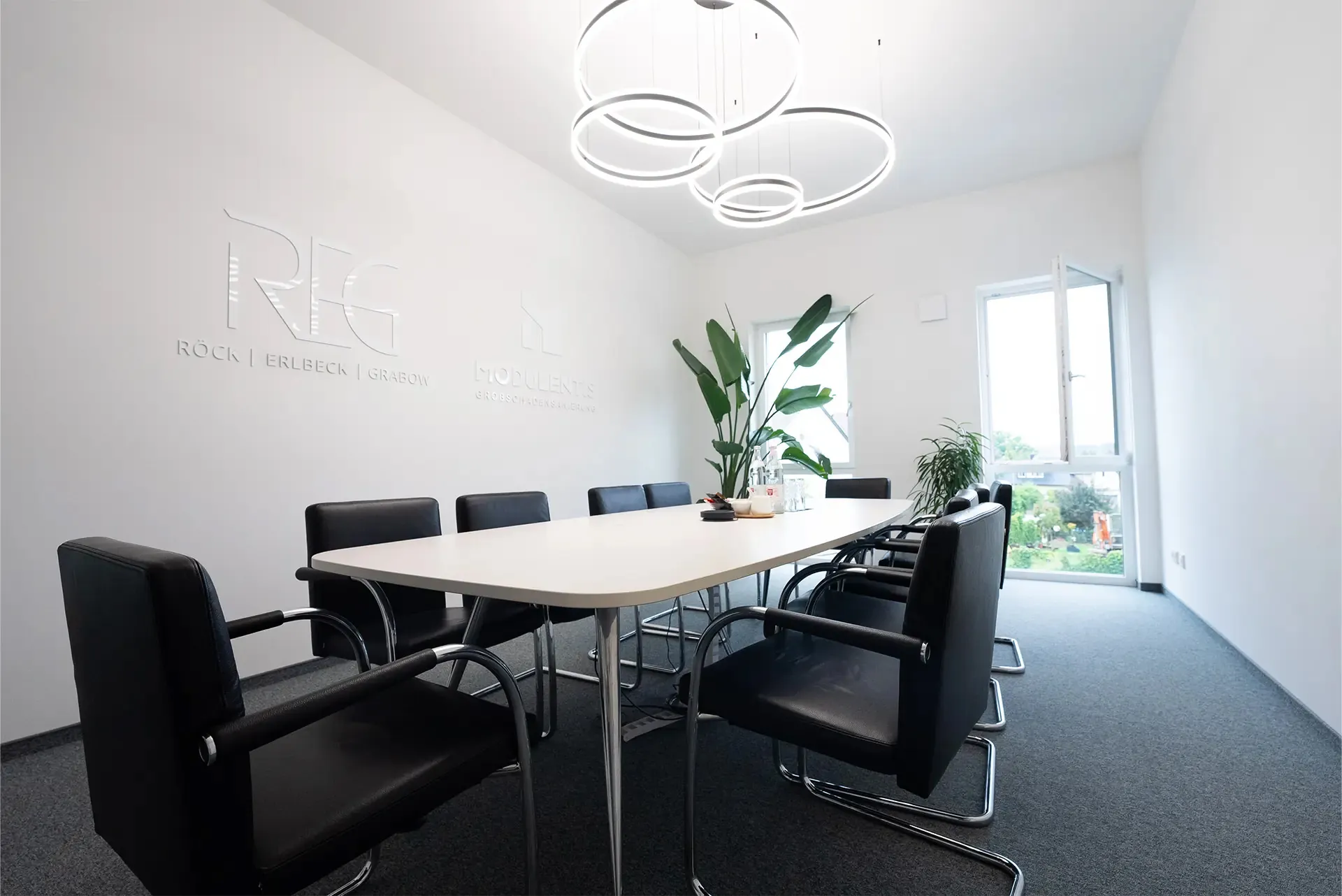 Besprechungsraum mit einem langen Tisch und zehn schwarzen Stühlen. An der Wand ist das Logo 'REG' in Reliefschrift. Eine große Pflanze steht neben dem Tisch, und zwei bodentiefe Fenster fluten den Raum mit Licht.