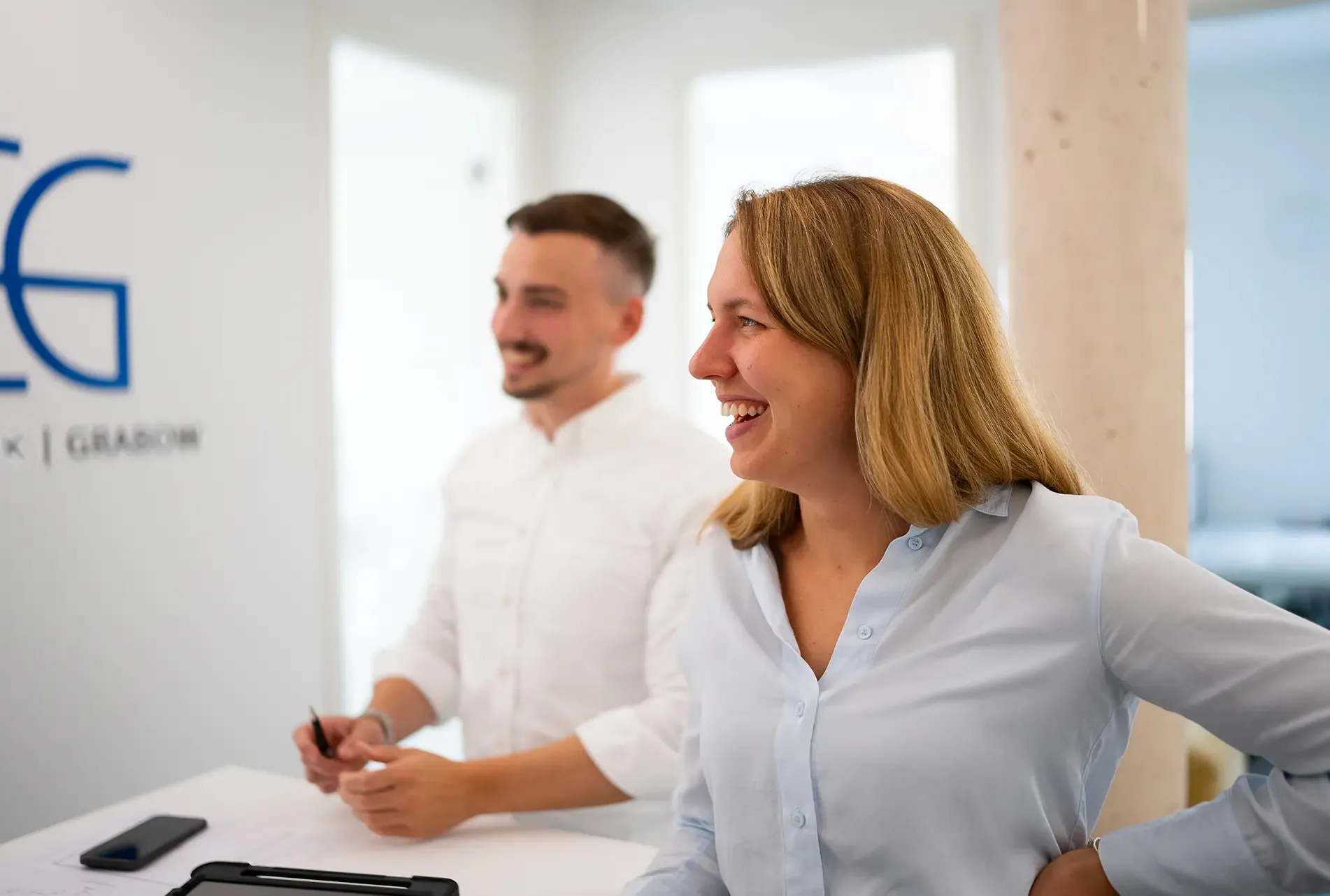 Zwei Kollegen im Büro lachen und arbeiten gemeinsam an einem Tisch in heller, moderner Arbeitsumgebung.
