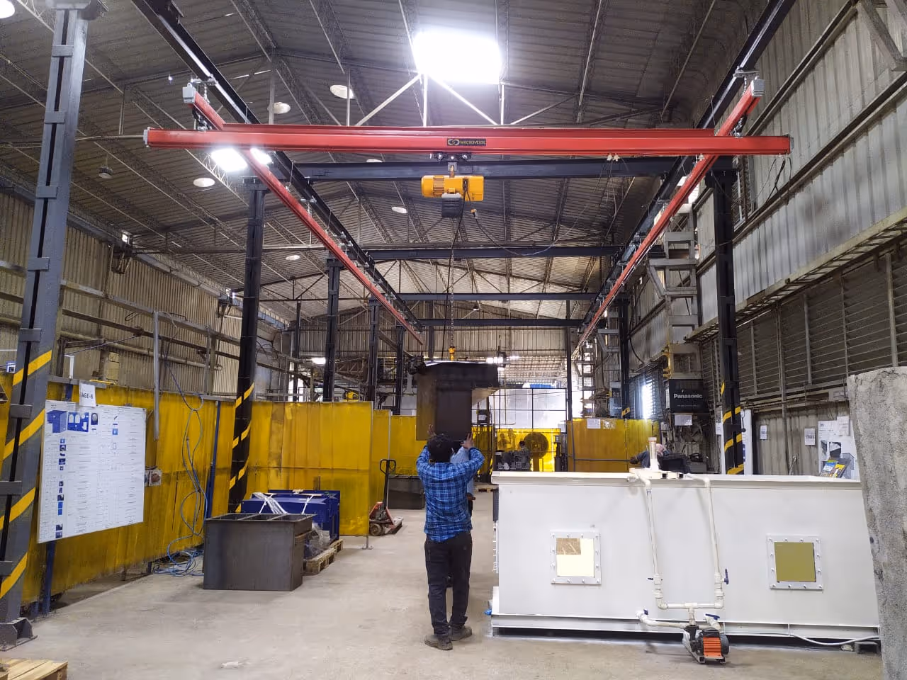 Two workers on blue lift inspecting a yellow overhead crane inside a large industrial warehouse.