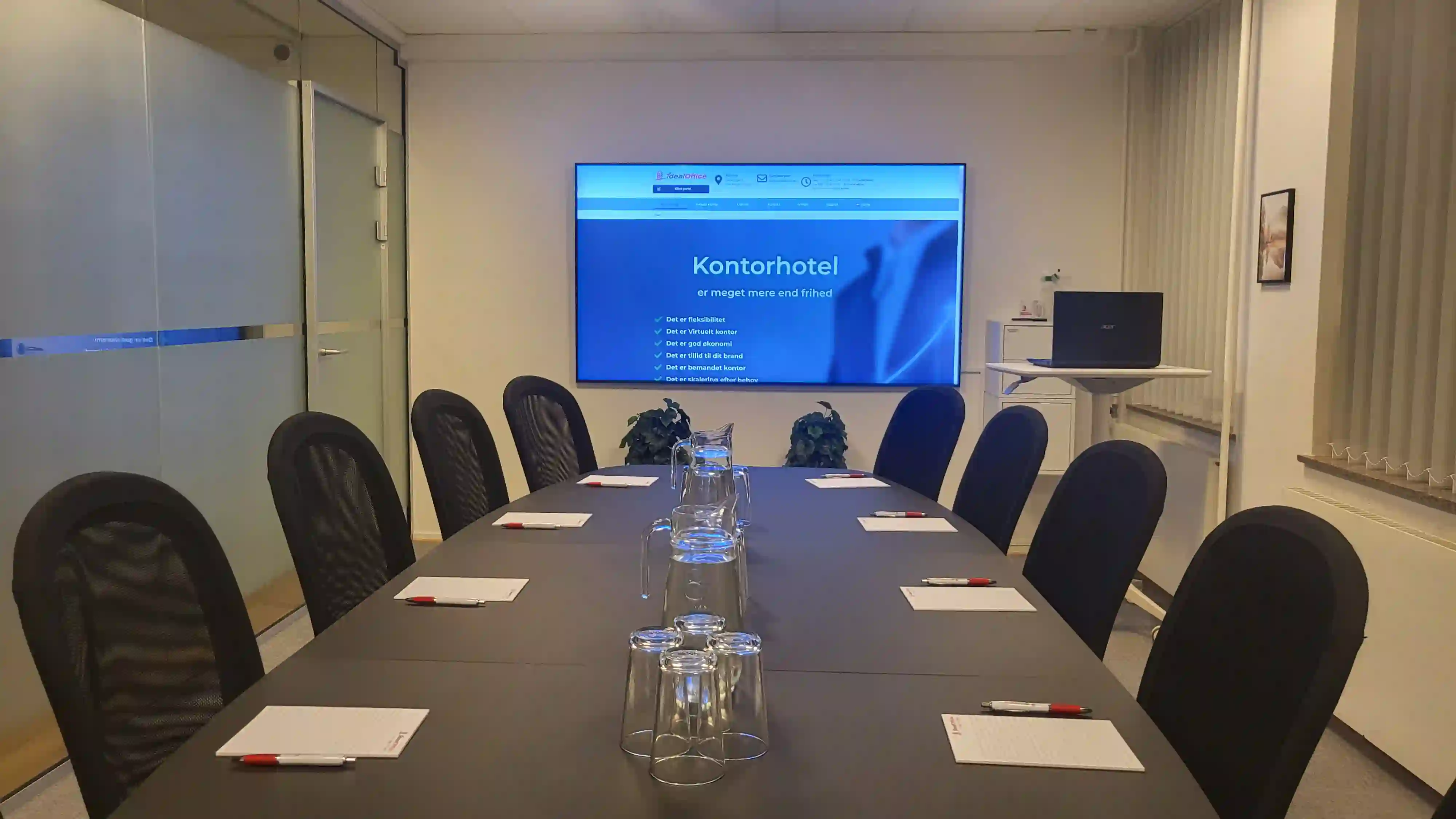 Moderne mødelokale med sort mødebord dækket op til møde med notesblokke, kuglepenne og glas. Skærm viser kontorhotel-information. To grønne planter står under skærmen.