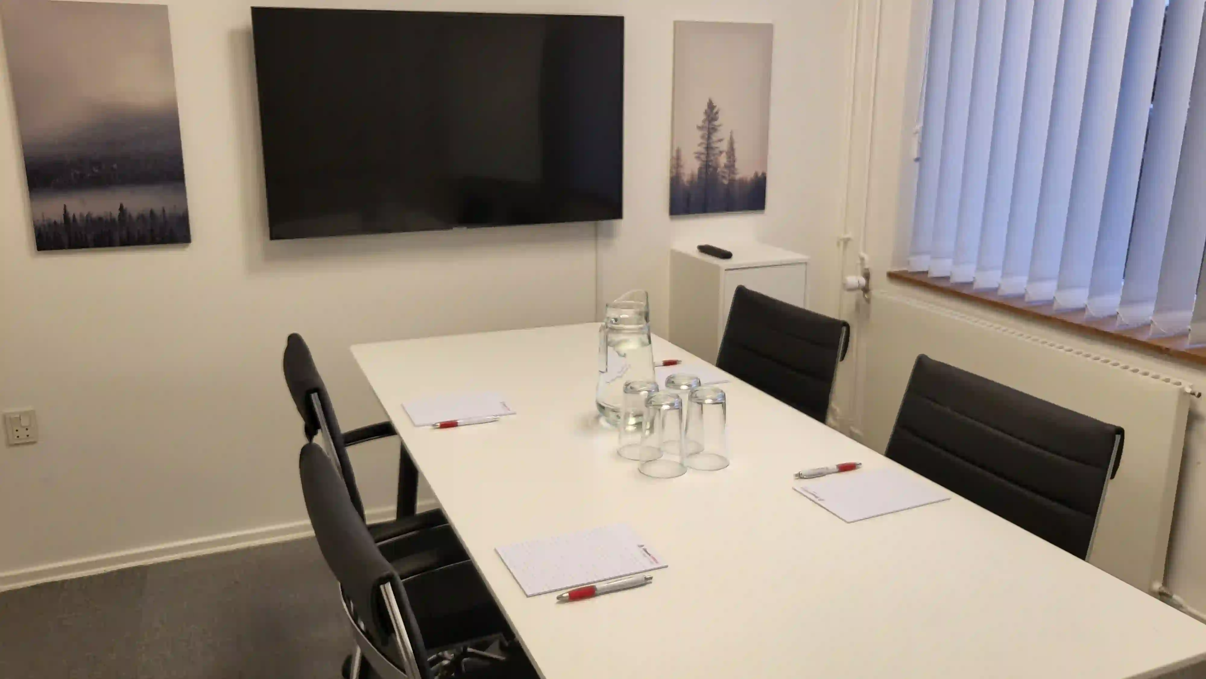 Mindre mødelokale med hvidt rektangulært bord dækket til fire personer. Rummet har sort lænestole, glas, vandkander og stor vægskærm omgivet af naturbilleder.