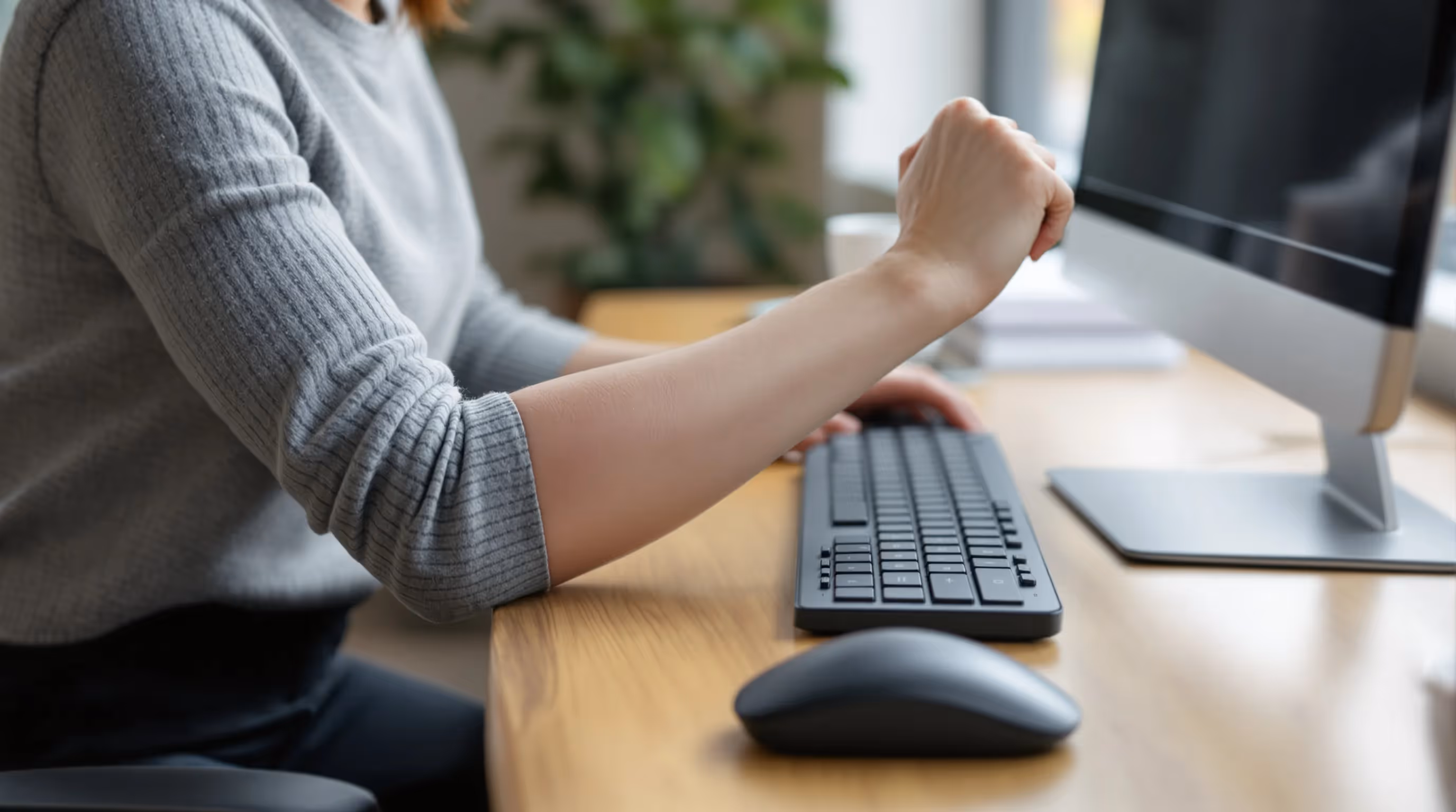 Woman at UK office desk showing outside elbow pain from far mouse reach.
