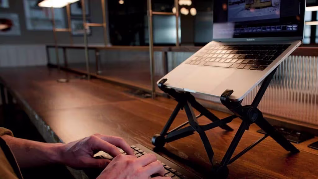 Portable adjustable laptop stand supporting elevated workstation ergonomics. Laptop positioned on a foldable stand above a desktop surface, creating a more ergonomic typing and viewing setup with an external keyboard.