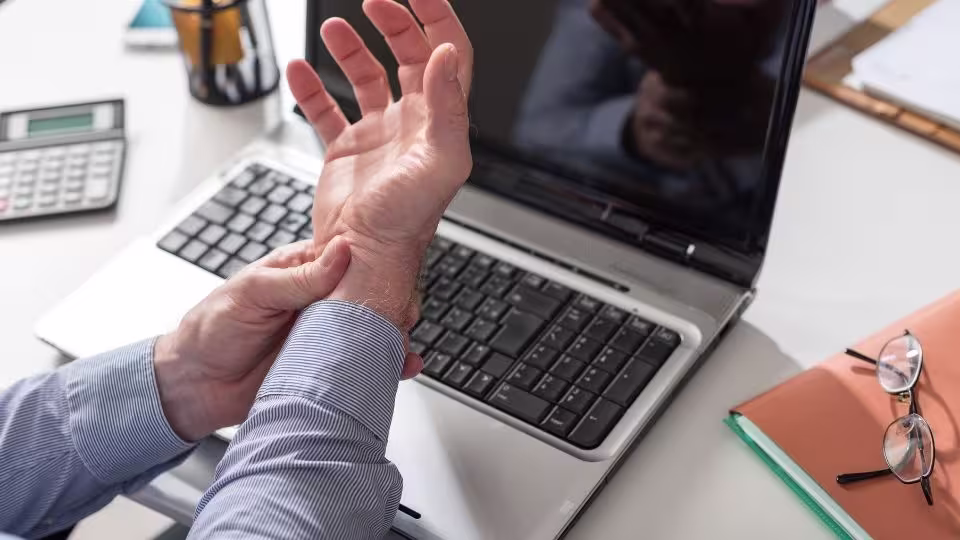 Henkilö, jolla on epämukava tunne ranteessa kannettavalla tietokoneella työskennellessä, mikä korostaa kyynärvarsituen tarvetta työpöydälle paremman ergonomisen ryhdin ja vähäisemmän rasituksen vuoksi.
