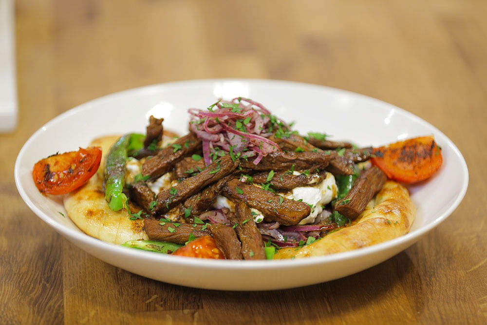 Tirit kebab in a wide white bowl grilled meat strips over pide bread and yogurt, topped with pickled red onion and chopped herbs