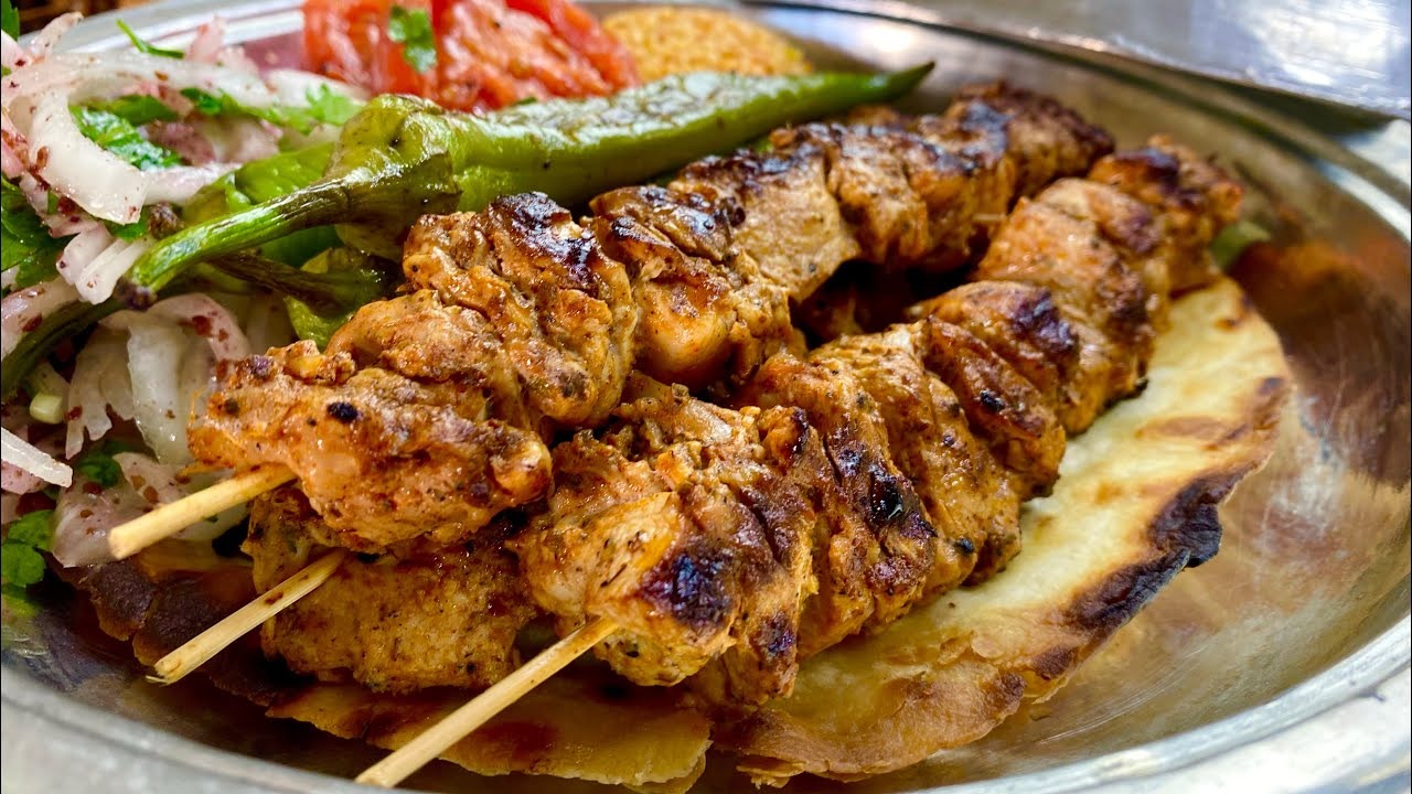 Close-up of grilled chicken shish kebab skewers served on flatbread with a grilled green pepper, a sliced onion and parsley salad, tomato, and rice, on a metal platter.